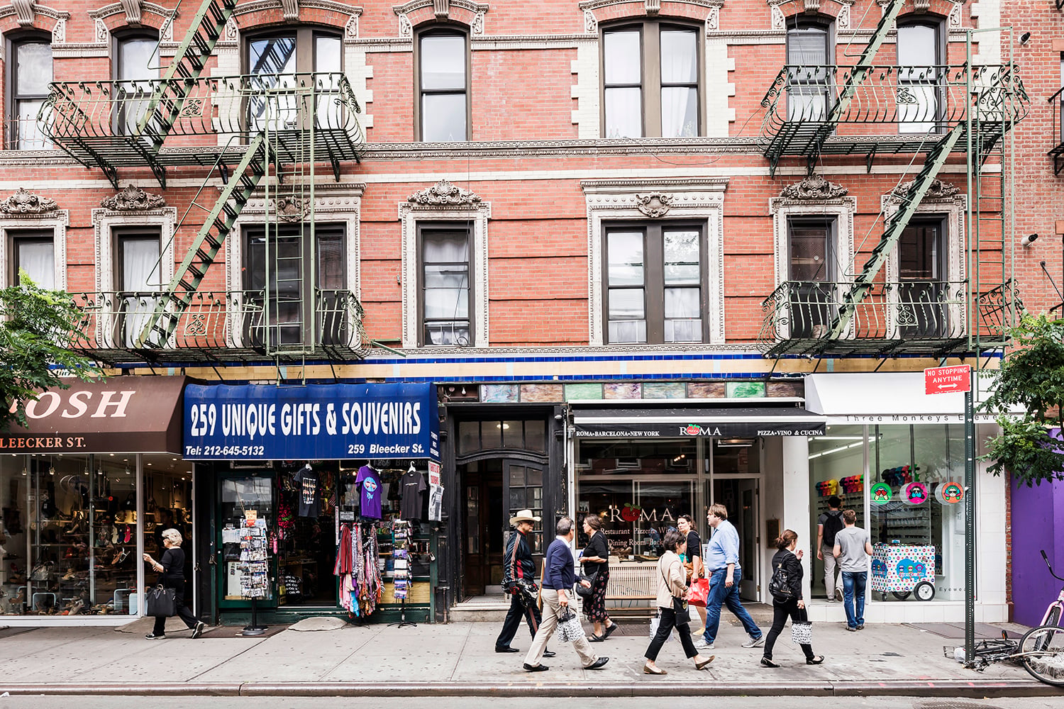 A group of people walk past storefronts, including a gift shop and a clothing store, on Bleecker Street with brick buildings and fire escapes.