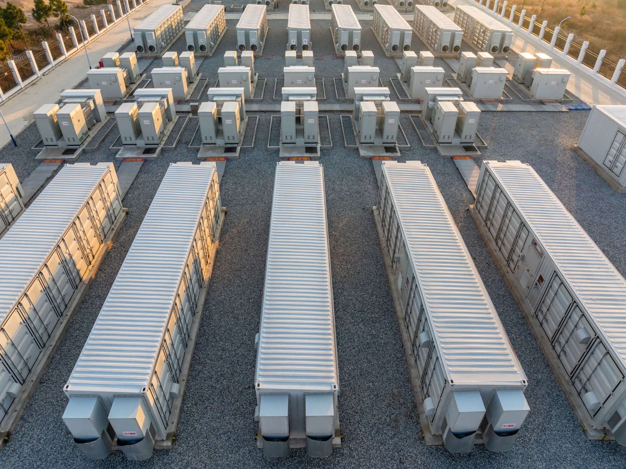 Aerial view of a large battery energy storage power station with rows of white storage containers and electrical equipment on a gravel lot, arranged in an organized grid pattern.