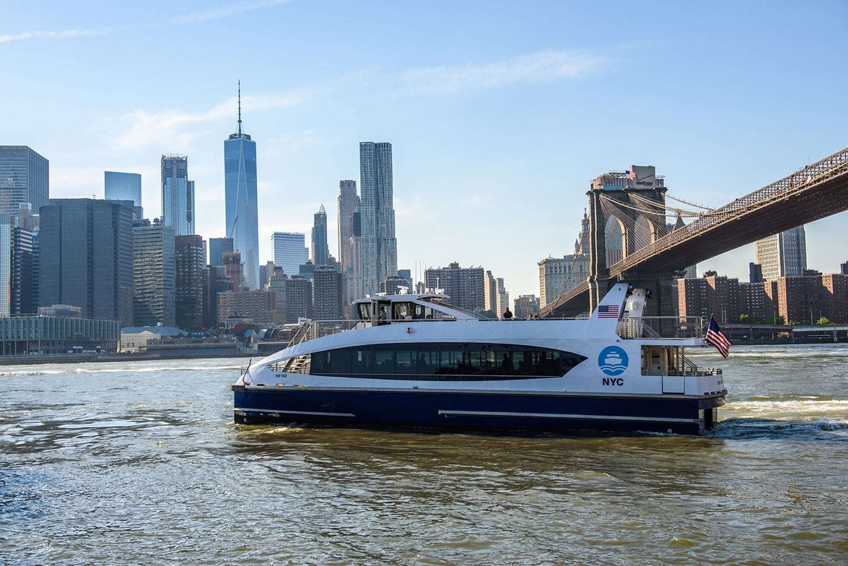 A white NYC ferry boat travels on the river with the Manhattan skyline and Brooklyn Bridge visible in the background on a clear day.