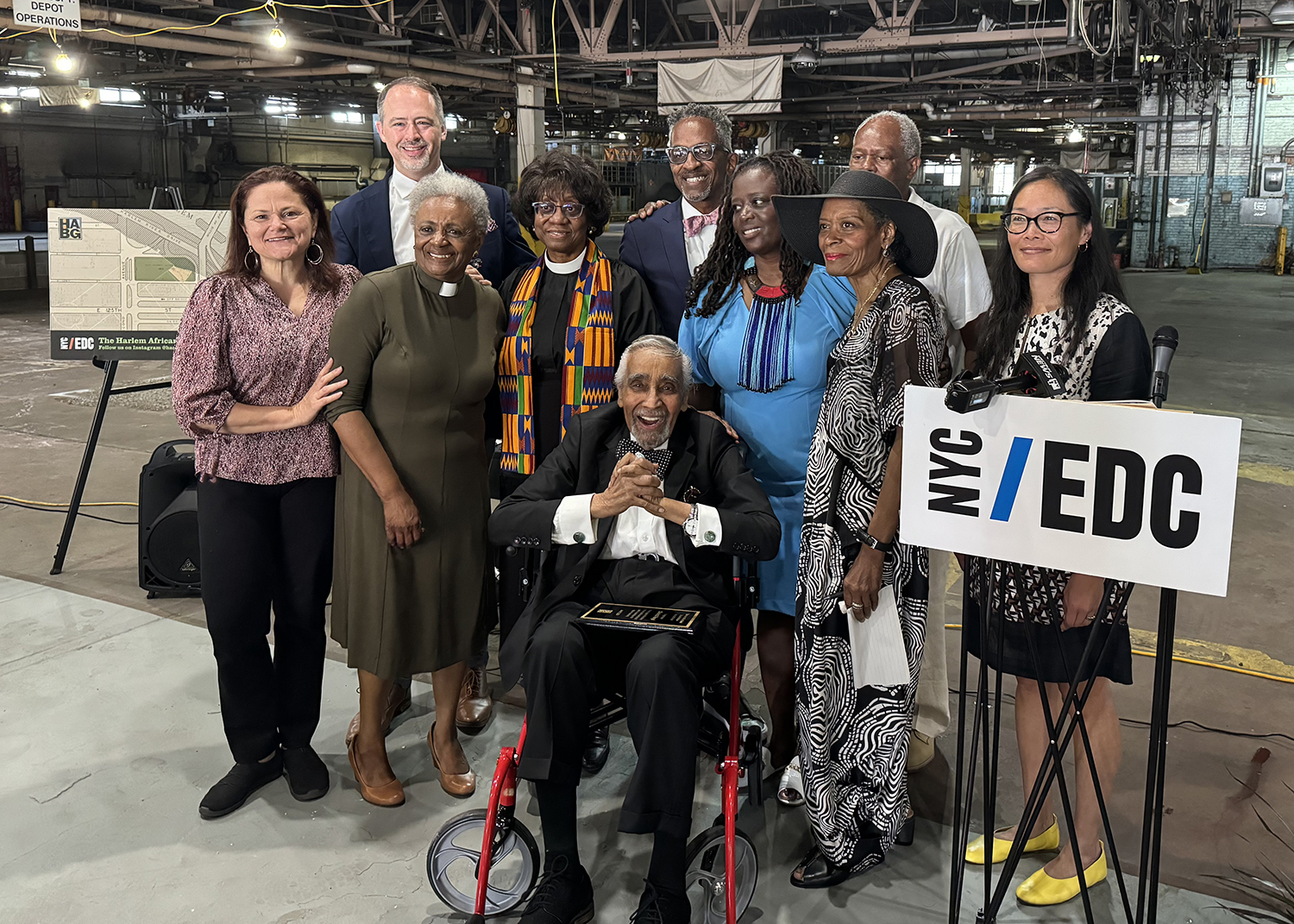 NYCEDC, Harlem African Burial Ground Initiative and Harlem Community members, including a man in a wheelchair and others standing, pose for a photo at the Harlem African Burial Ground beside a podium with a sign that reads “NYC/EDC.” All are dressed in formal or business attire.