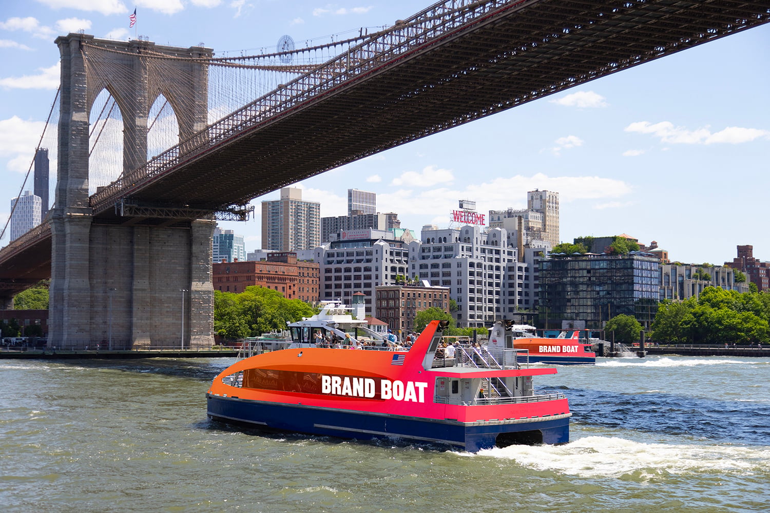 An orange and blue ferry labeled "BRAND BOAT" cruises under the Brooklyn Bridge with the New York City skyline in the background on a sunny day.