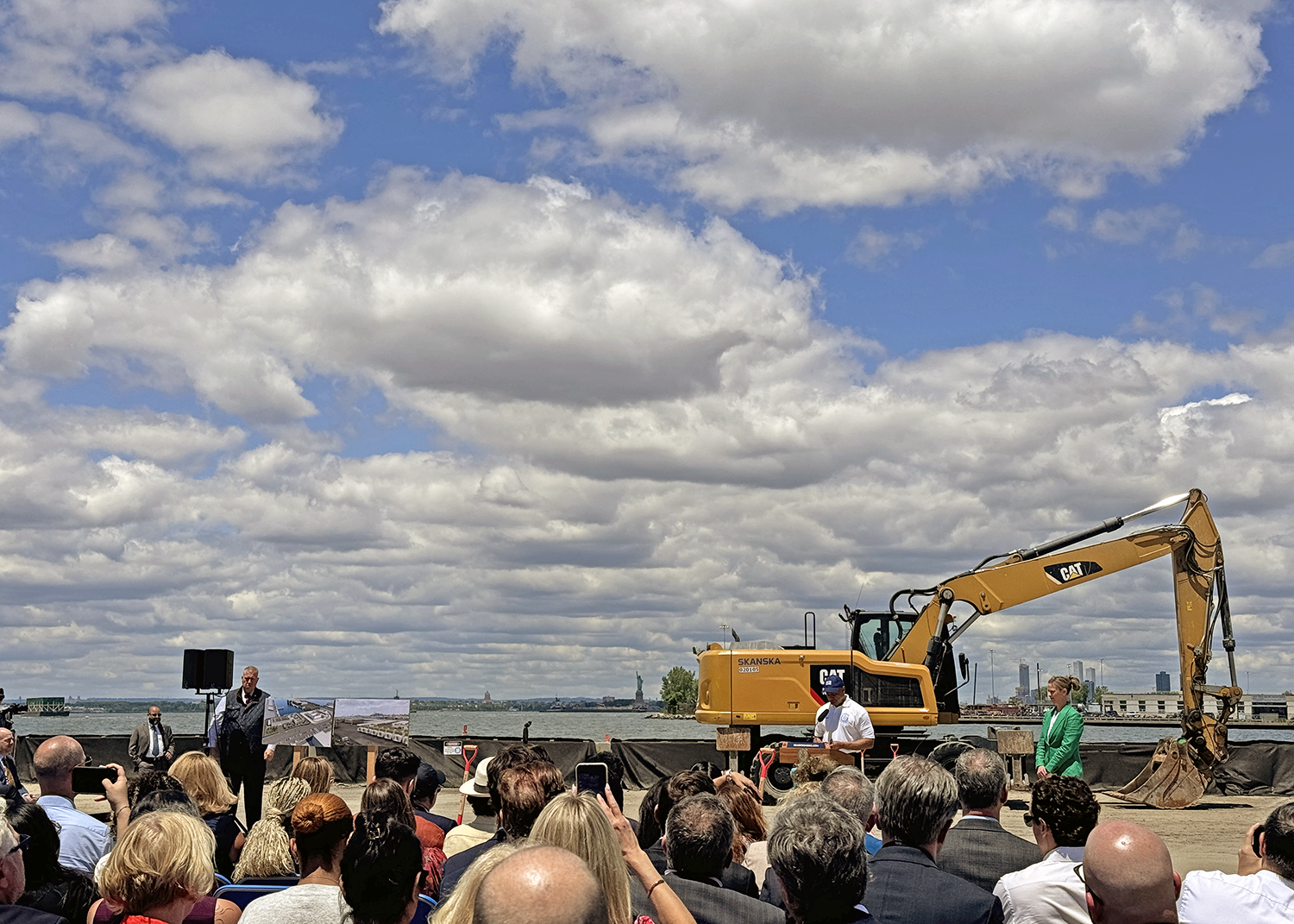 A group of people listening to NYC Mayor Eric Adams speaking at the South Brooklyn Marine Terminal groundbreaking ceremony with a large excavator in the background and a partly cloudy sky.