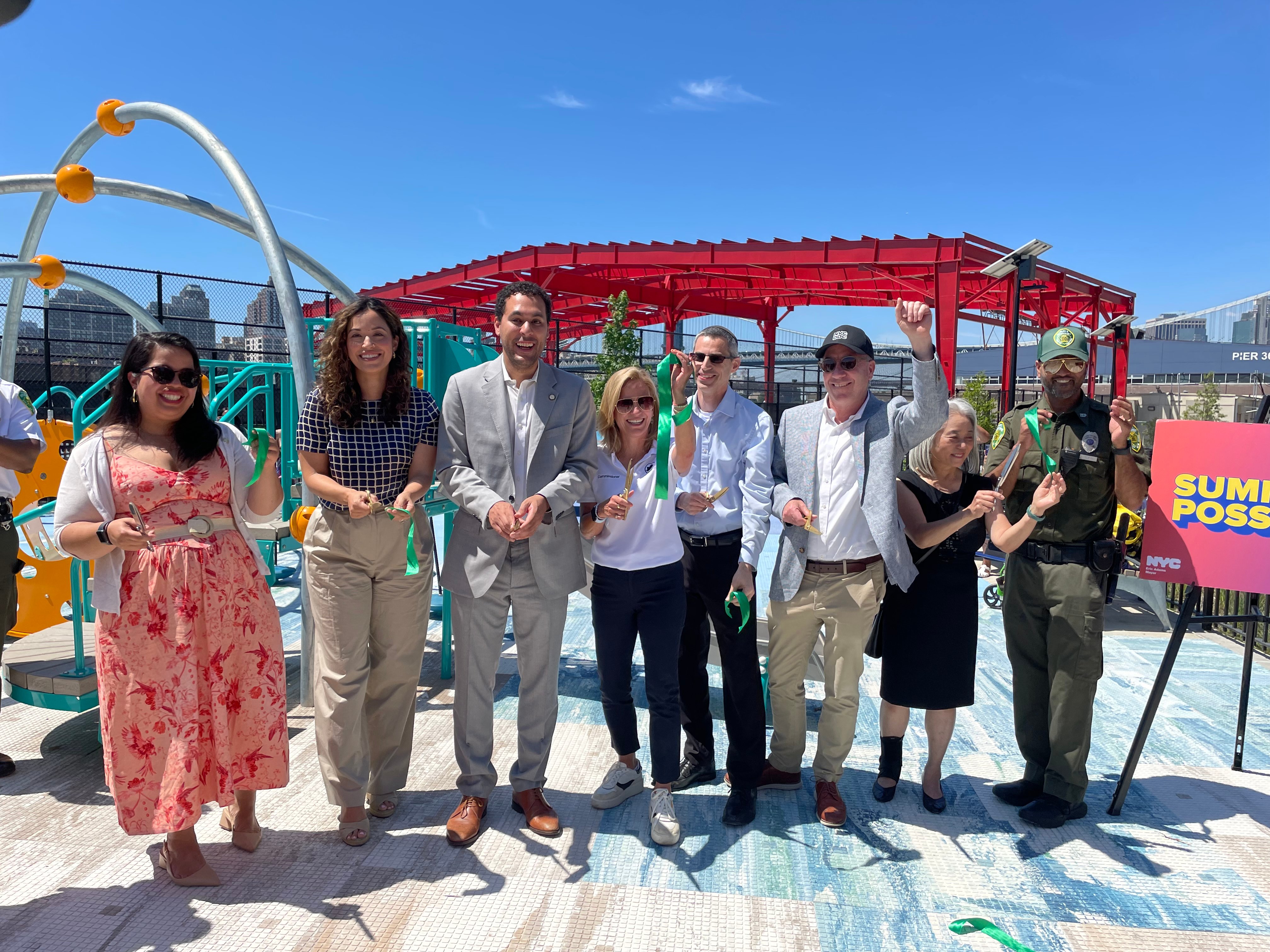 A group of eight New York City Officials stand in a row and hold pieces of a green ribbon at a playground to celebrate the opening of Pier 42 open space.