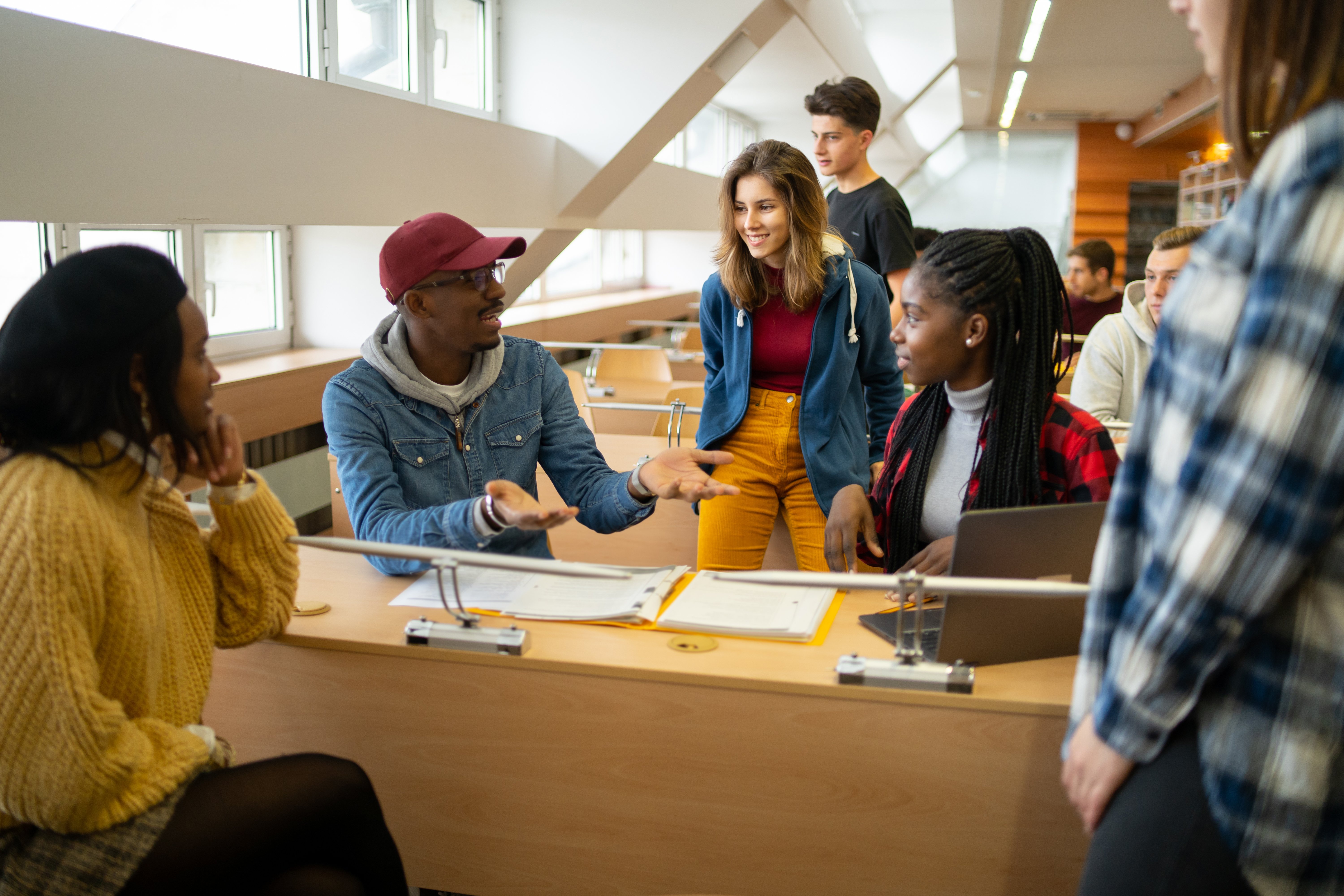 A group of students in a classroom engage in discussion, sitting and standing around desks with papers and a laptop.