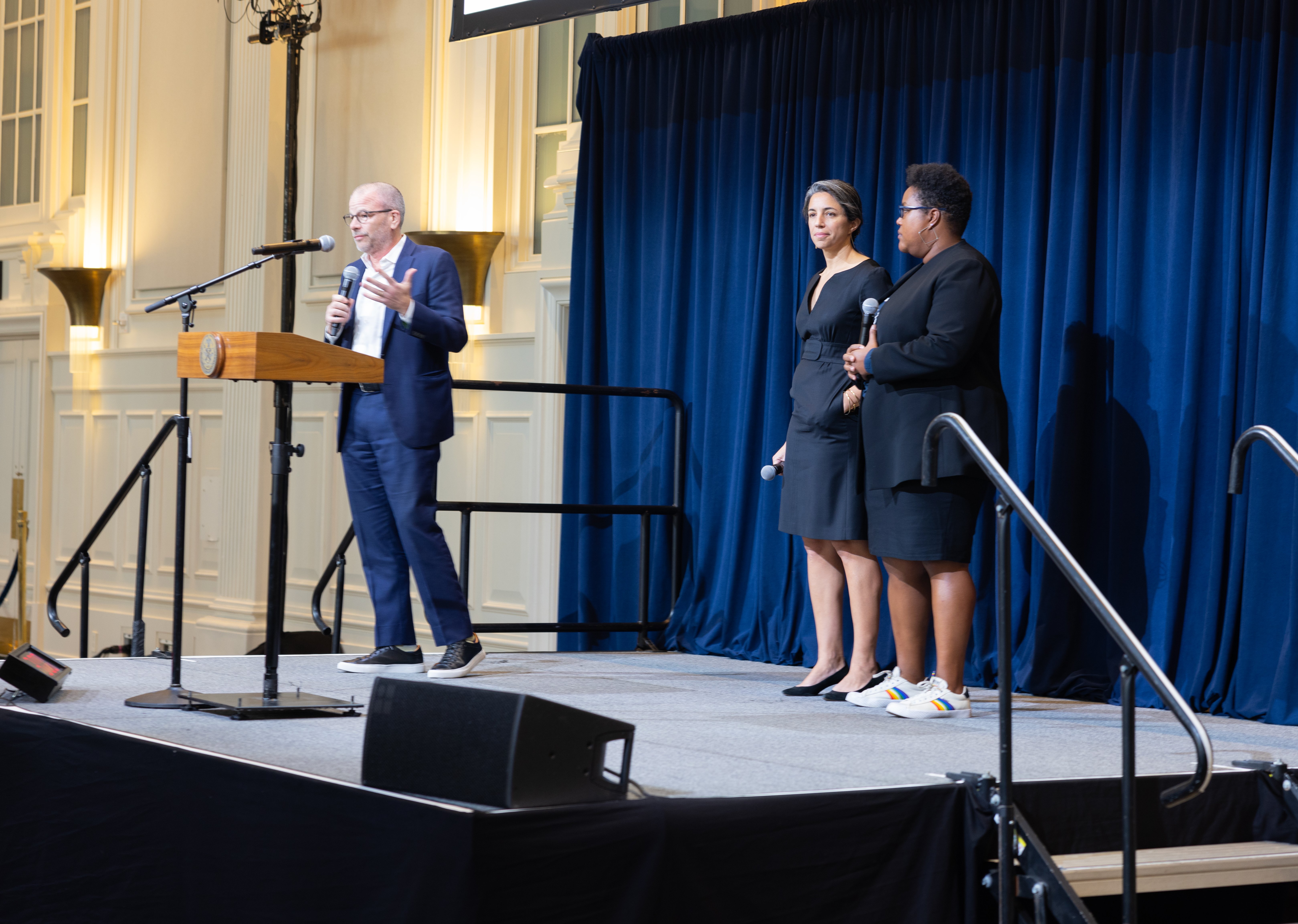 Three people standing on a stage at the NYC Climate Technology Showcase; Andrew Kimball is speaking at a podium, while the other two hold microphones.