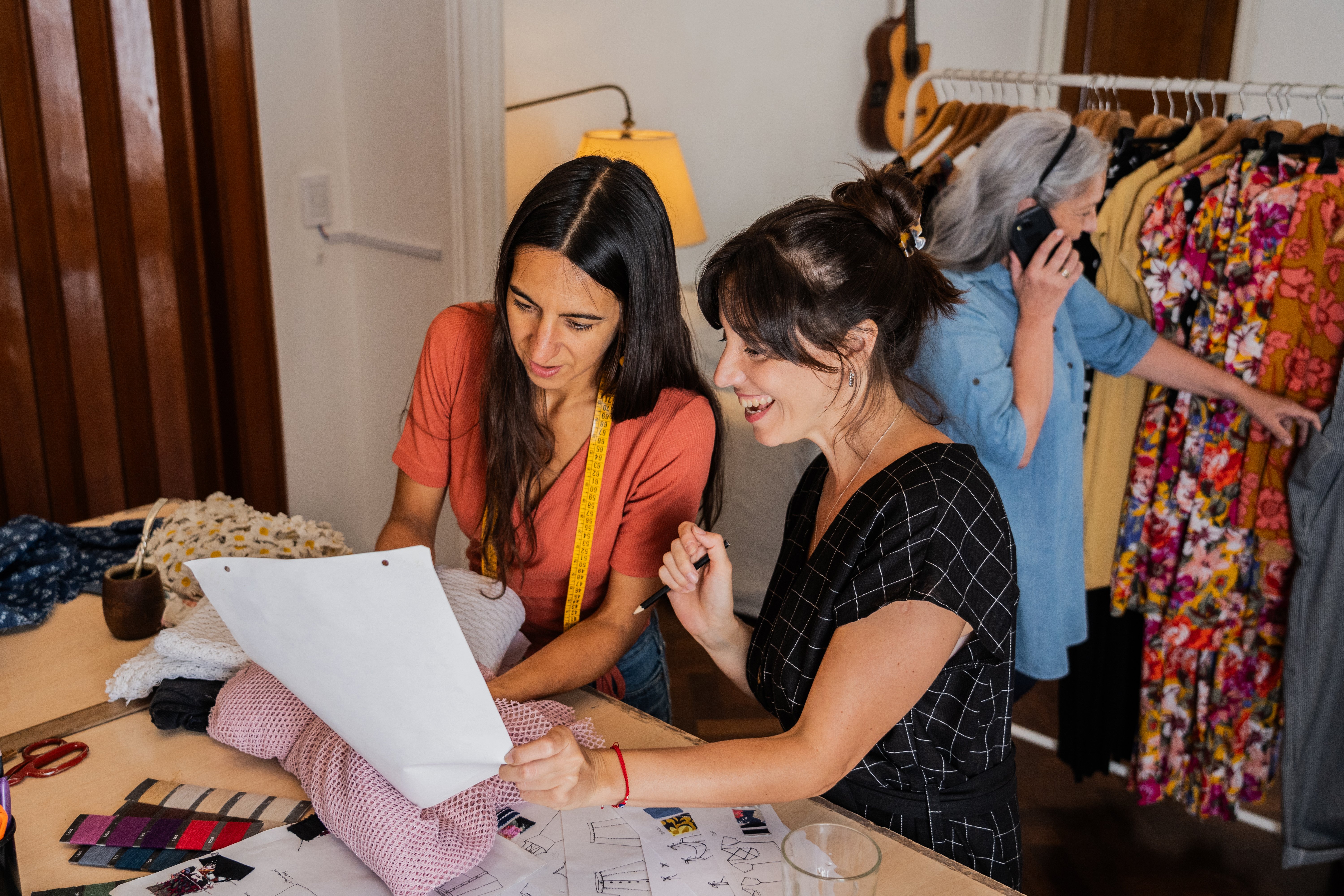 Two women fashion designers are looking at a sheet of paper and fabric samples in a sewing room. Another woman in the background is talking on the phone by a clothing rack.
