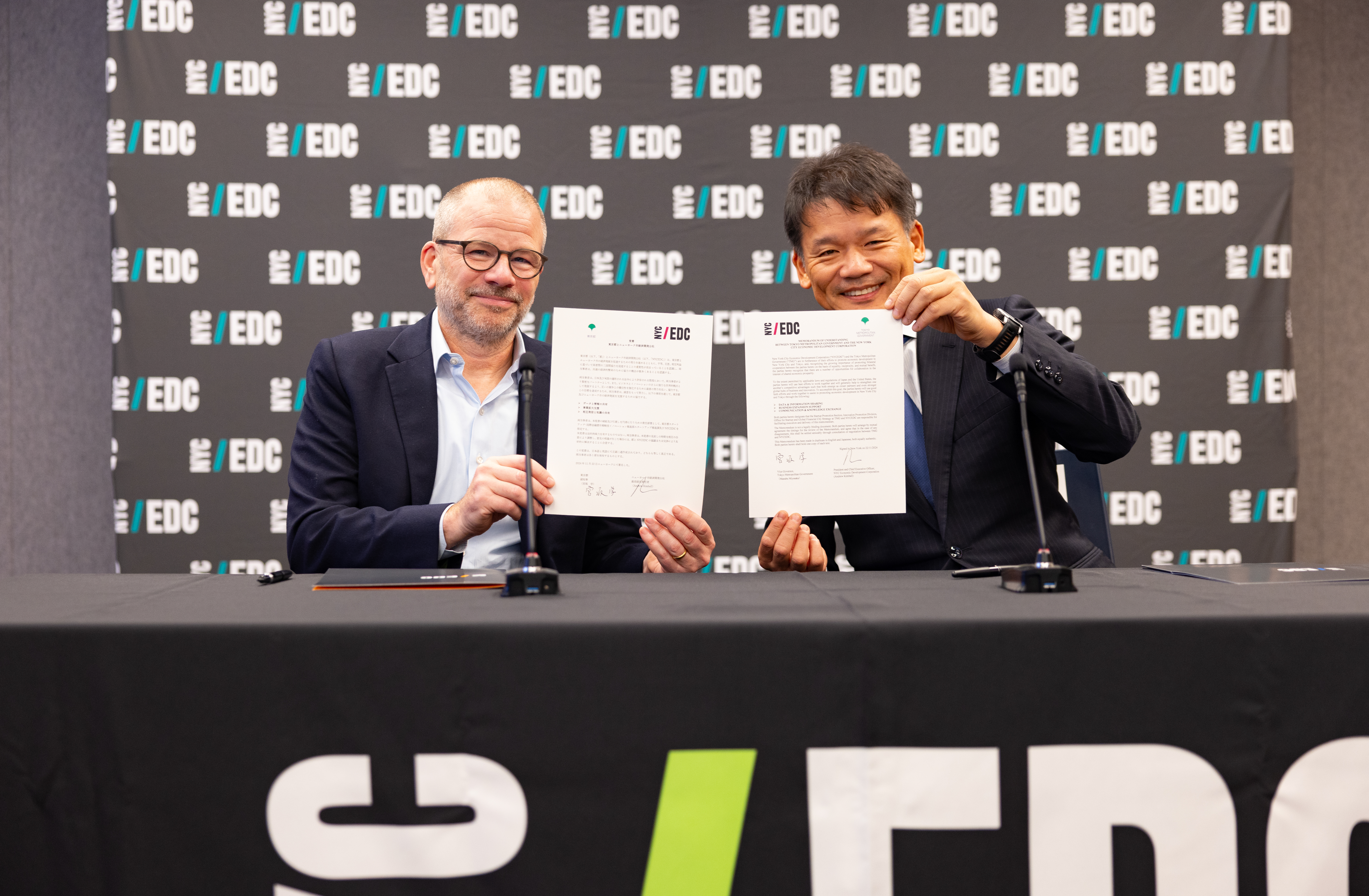 Two men seated at a table hold up signed MOU documents. A banner behind them displays the EDC logo repeatedly.