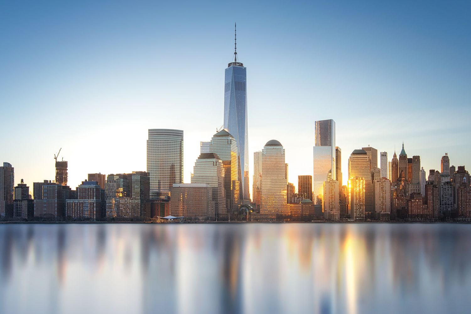 Skyline of New York City with modern high-rise buildings reflecting in calm water, under a clear blue sky at sunset.