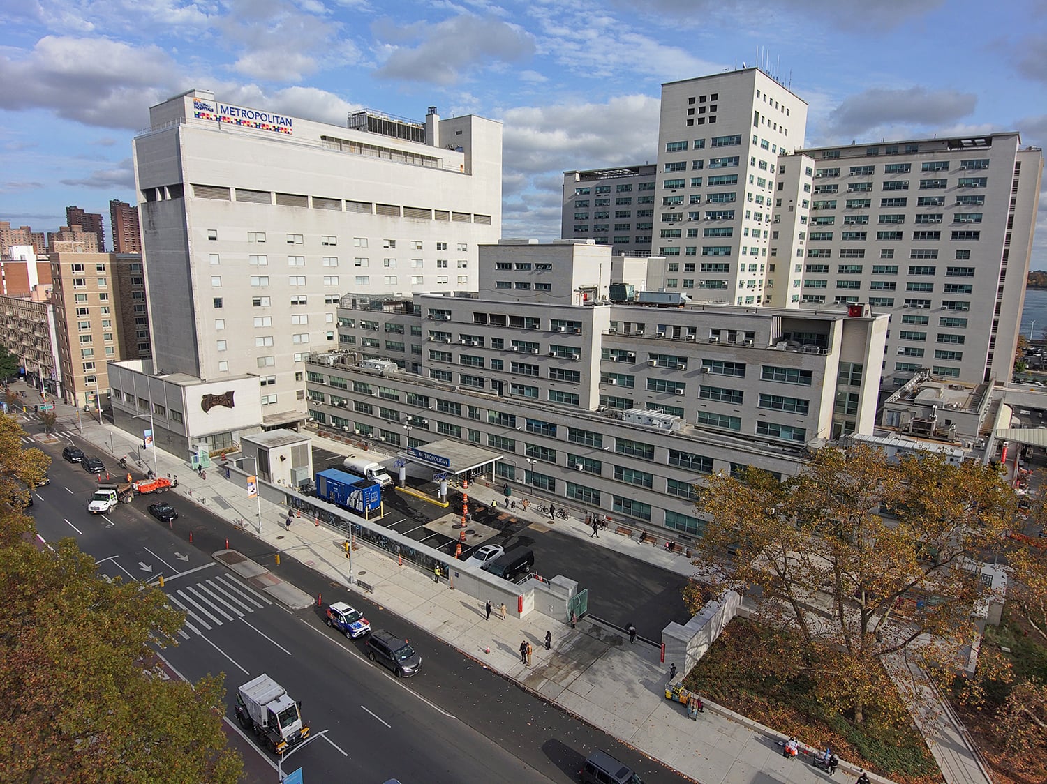 Aerial view of the large Metropolitan Hospital complex with multiple buildings and a busy street in the foreground. Trees with autumn foliage line the area.