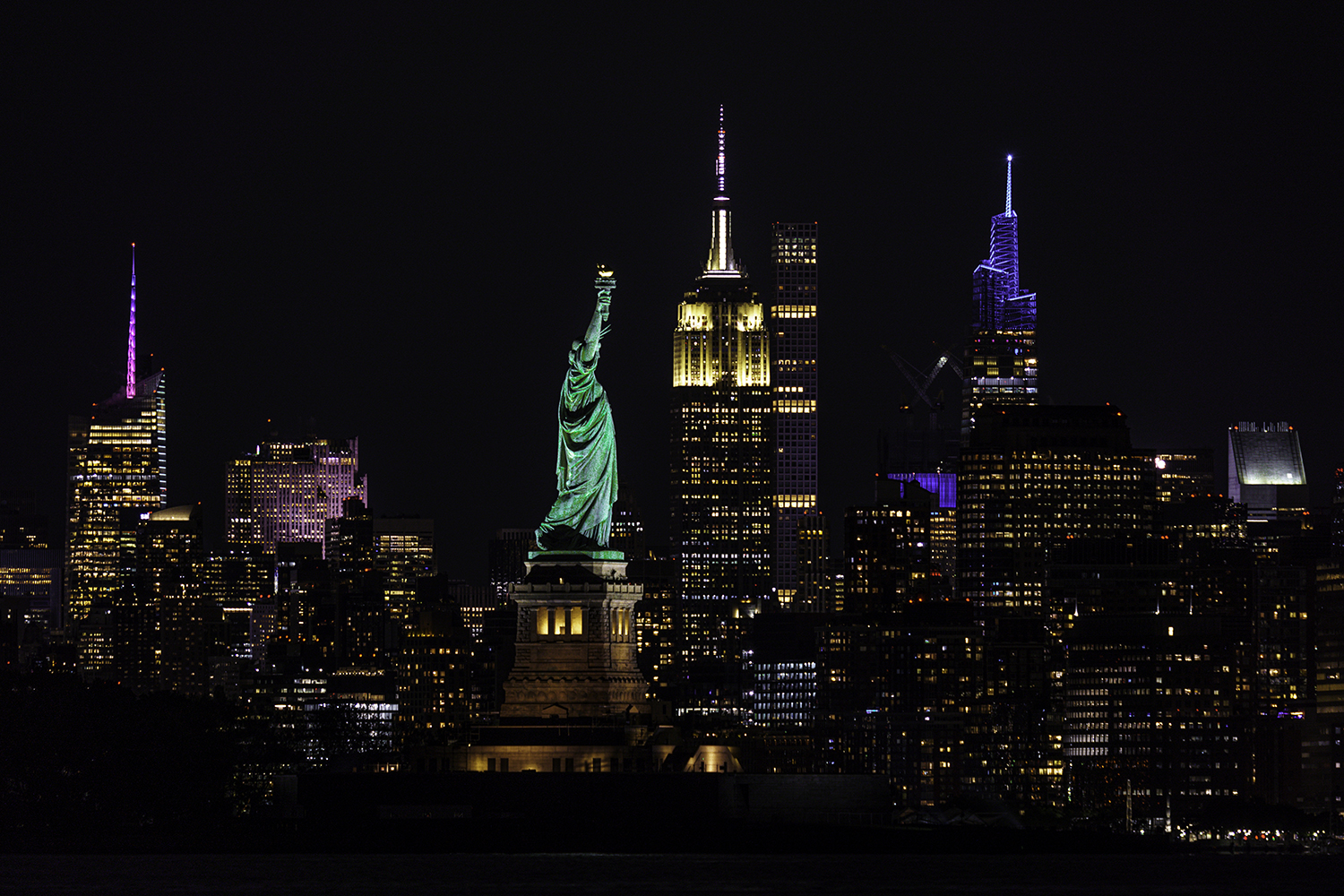 Night view of the New York City skyline featuring the illuminated Statue of Liberty in the foreground and skyscrapers in the background.