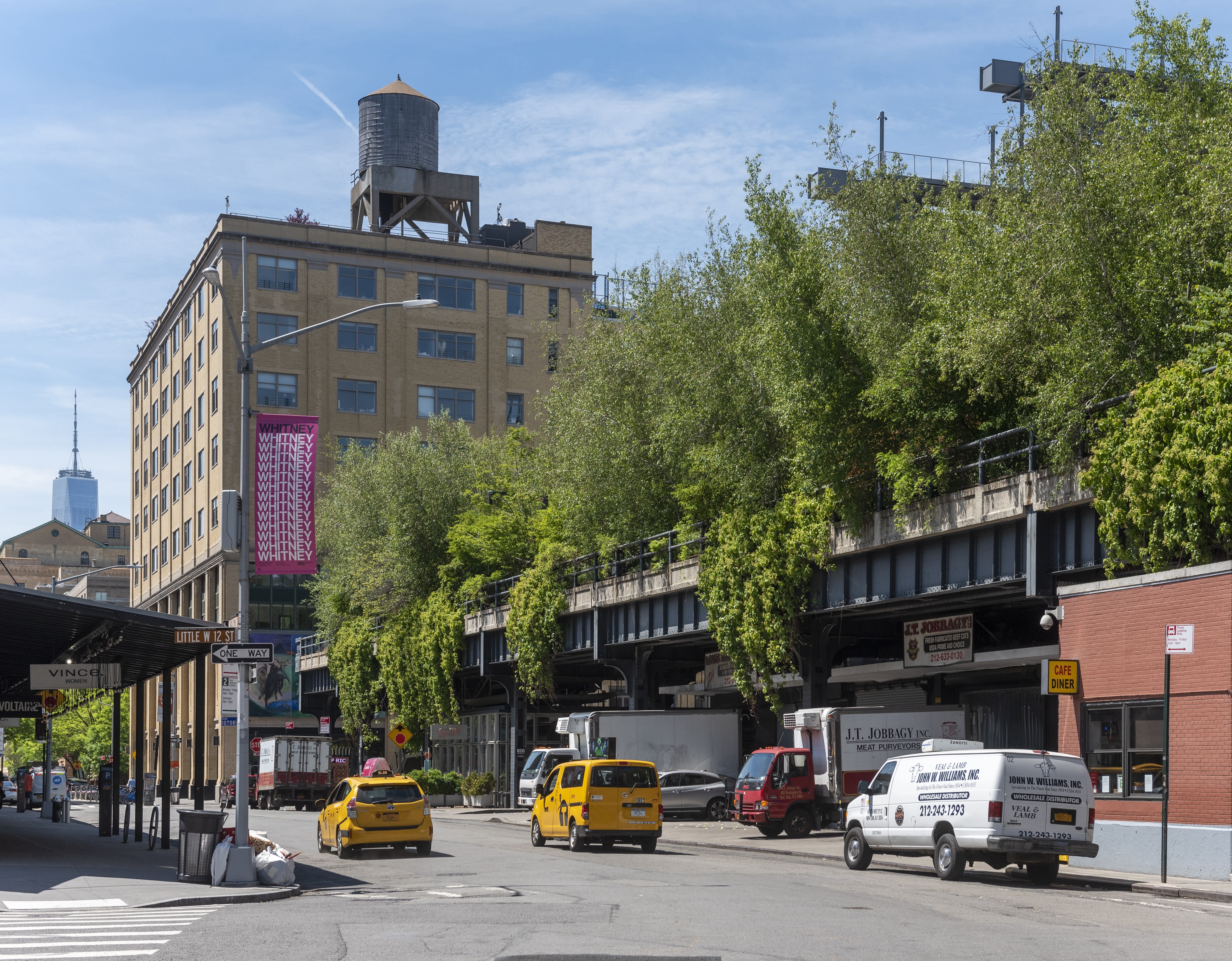 Street scene with a view of the High Line in New York City. The old railway track is lined with greenery and bordered by buildings. Yellow taxis, a van, and a café with a red awning are visible. The One World Trade Center is in the background.
