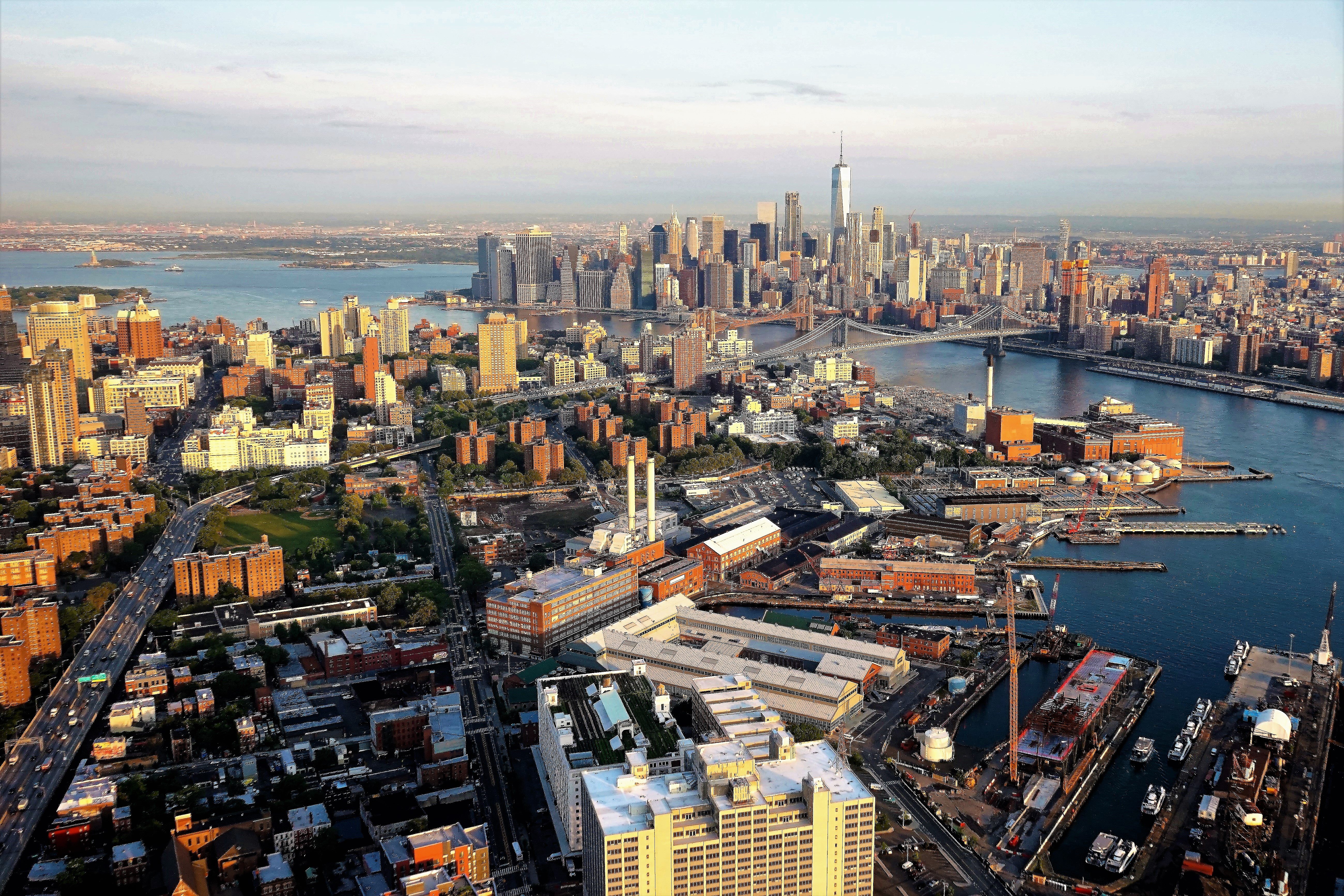 Aerial view of Manhattan, New York City, showcasing skyscrapers, the Hudson River, and surrounding boroughs. The One World Trade Center is prominently visible, with sunlit buildings and bustling waterways in the foreground.