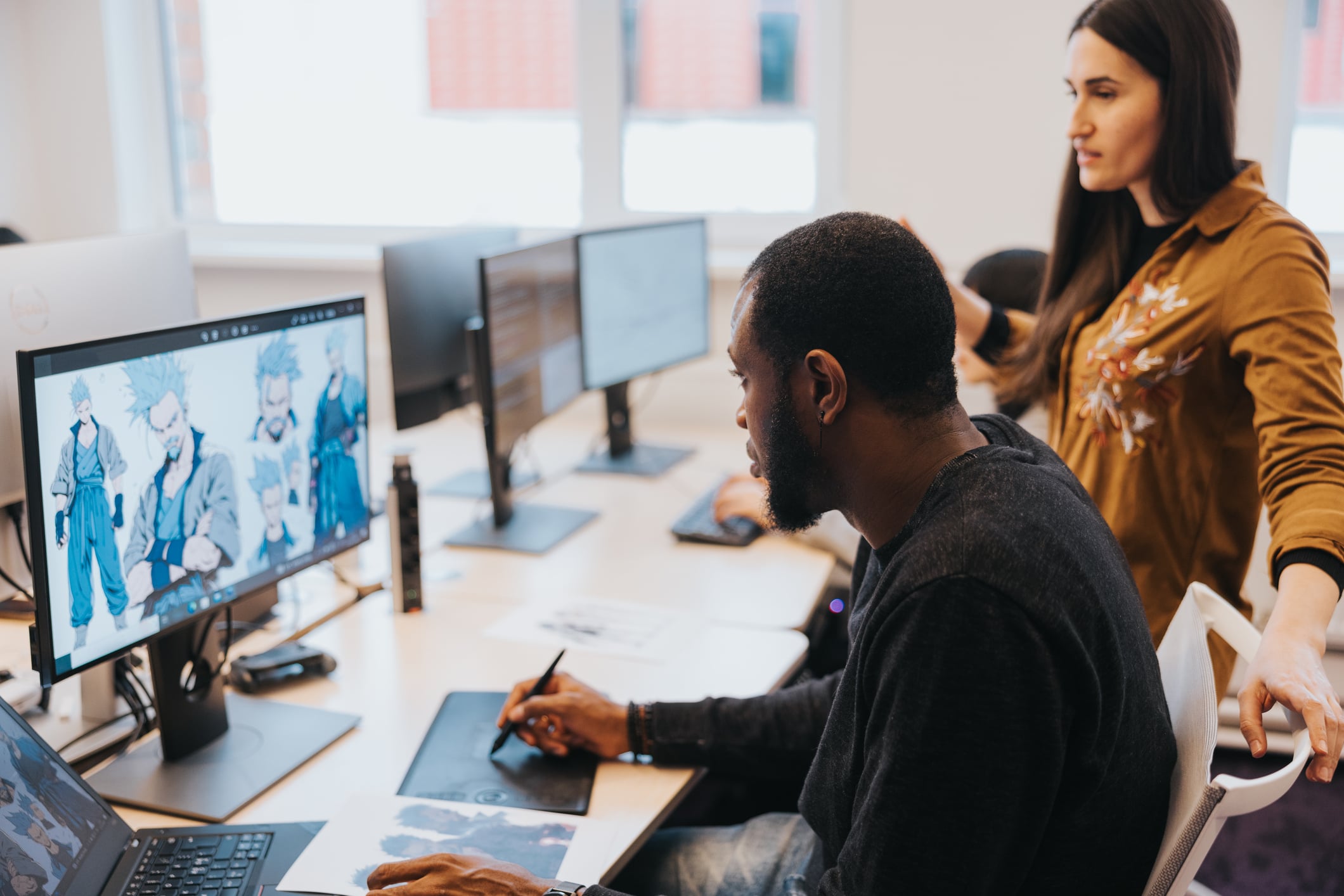 A man uses a drawing tablet and stylus while working on a character design on a computer screen in an office. A woman stands beside him, looking at the screen. Other monitors and desks are visible in the background.