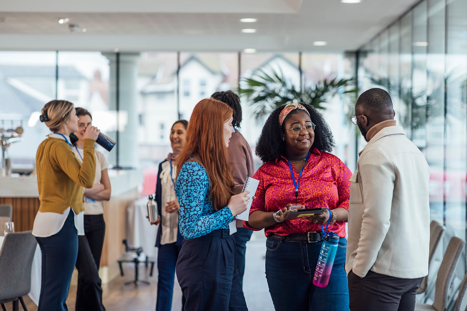 A diverse group of people stand in a modern office space, engaged in conversation. Some hold water bottles and papers. Large windows and soft lighting create an open, airy atmosphere.