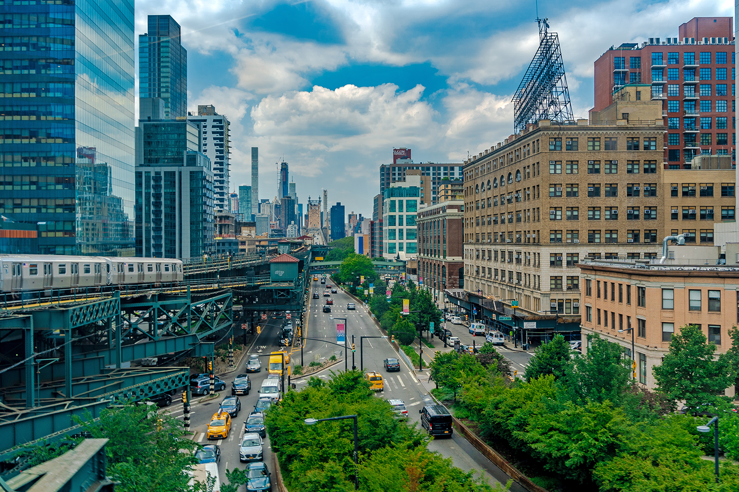 A busy urban street with cars and yellow cabs runs between tall buildings. A subway train travels on an elevated track to the left. The New York City skyline is visible in the background under a partly cloudy sky.