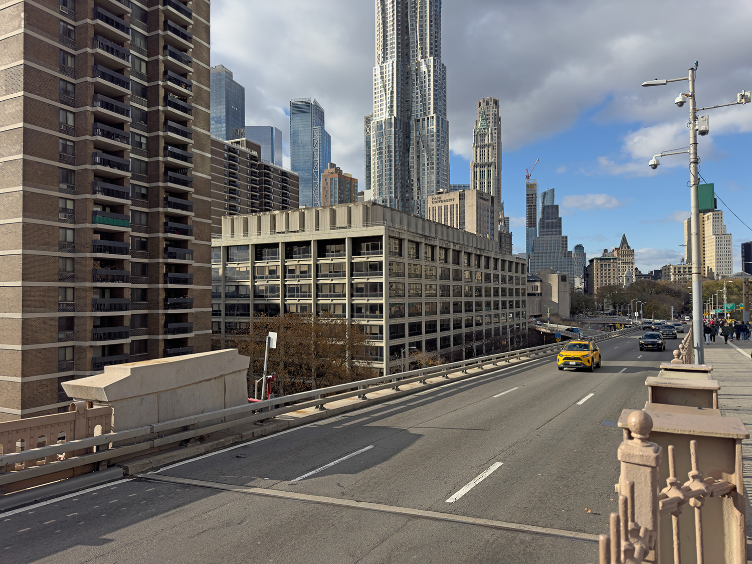 Urban scene at 100 Gold Street with a yellow taxi driving on a bridge in Manhattan. Tall skyscrapers are in the background beneath a partly cloudy sky. The setting appears busy and modern with multiple lanes on the road.