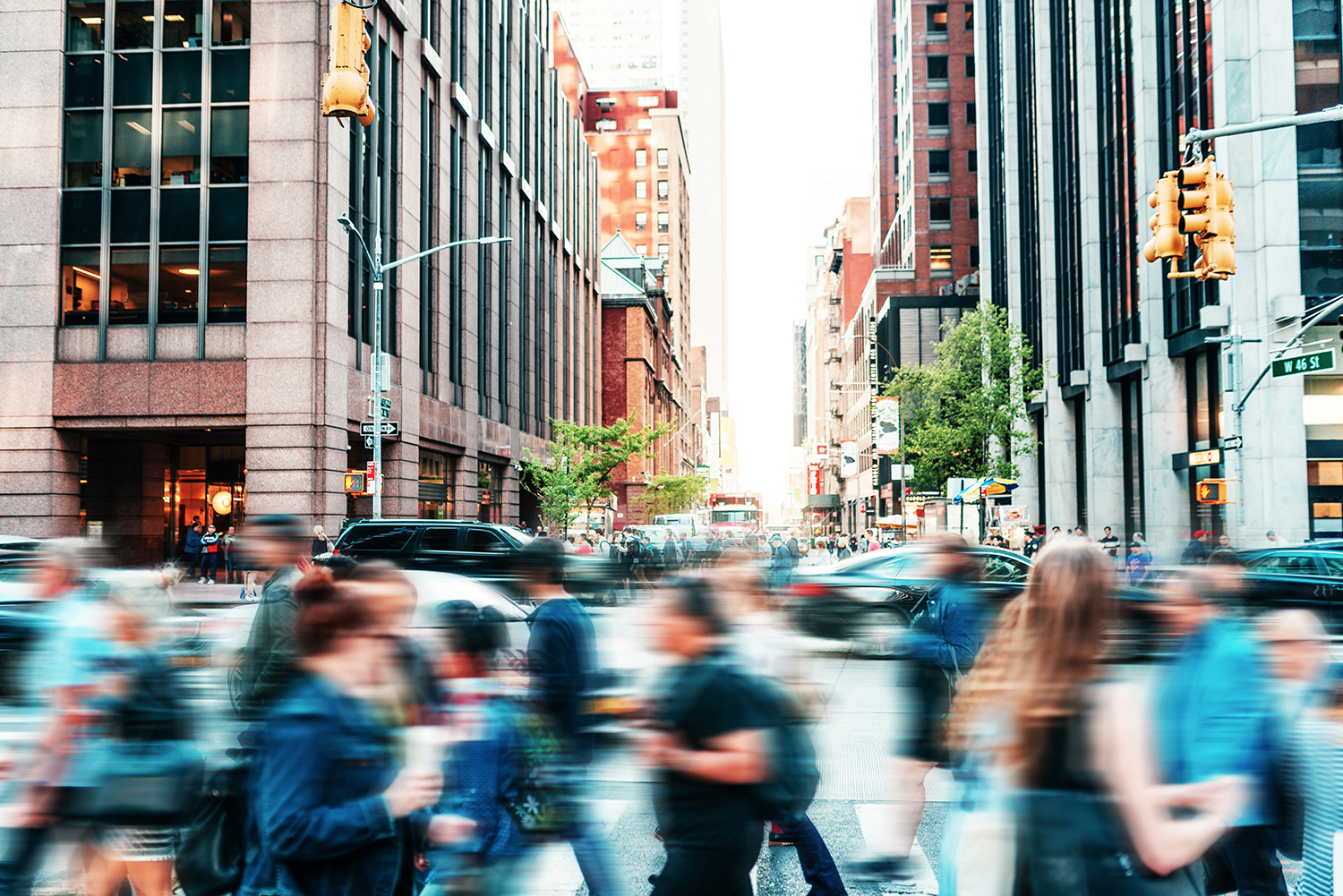 A busy New York City street scene with blurred motion capturing people crossing at a crosswalk. Tall buildings line both sides of the street, and the background reveals a mix of trees and urban architecture. Traffic lights are visible above the intersection.