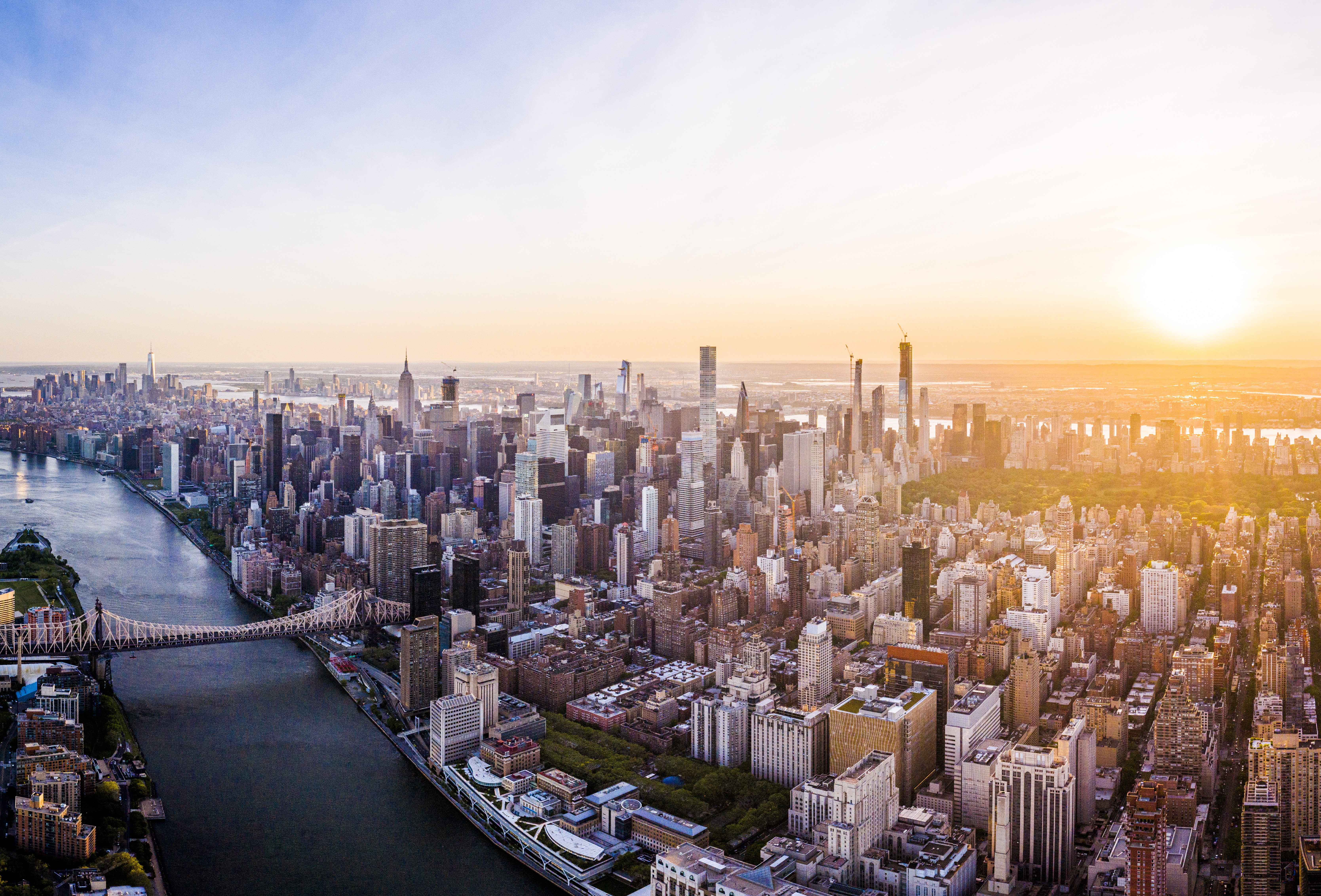 Aerial view of New York City skyline during sunset, featuring numerous skyscrapers and buildings. A wide river runs alongside the city, crossed by a prominent bridge. The sky is clear with the sun setting on the horizon.
