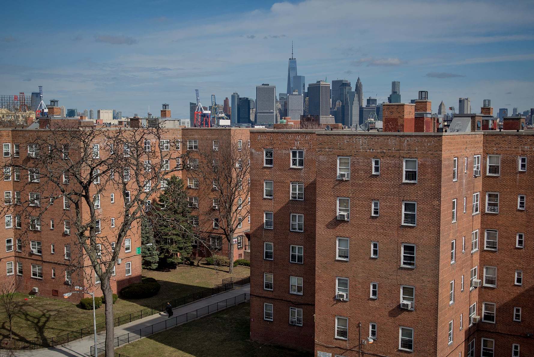 Red brick apartment buildings with small lawns and leafless trees in the foreground; the New York City skyline with skyscrapers and the One World Trade Center is visible in the background under a partly cloudy sky.