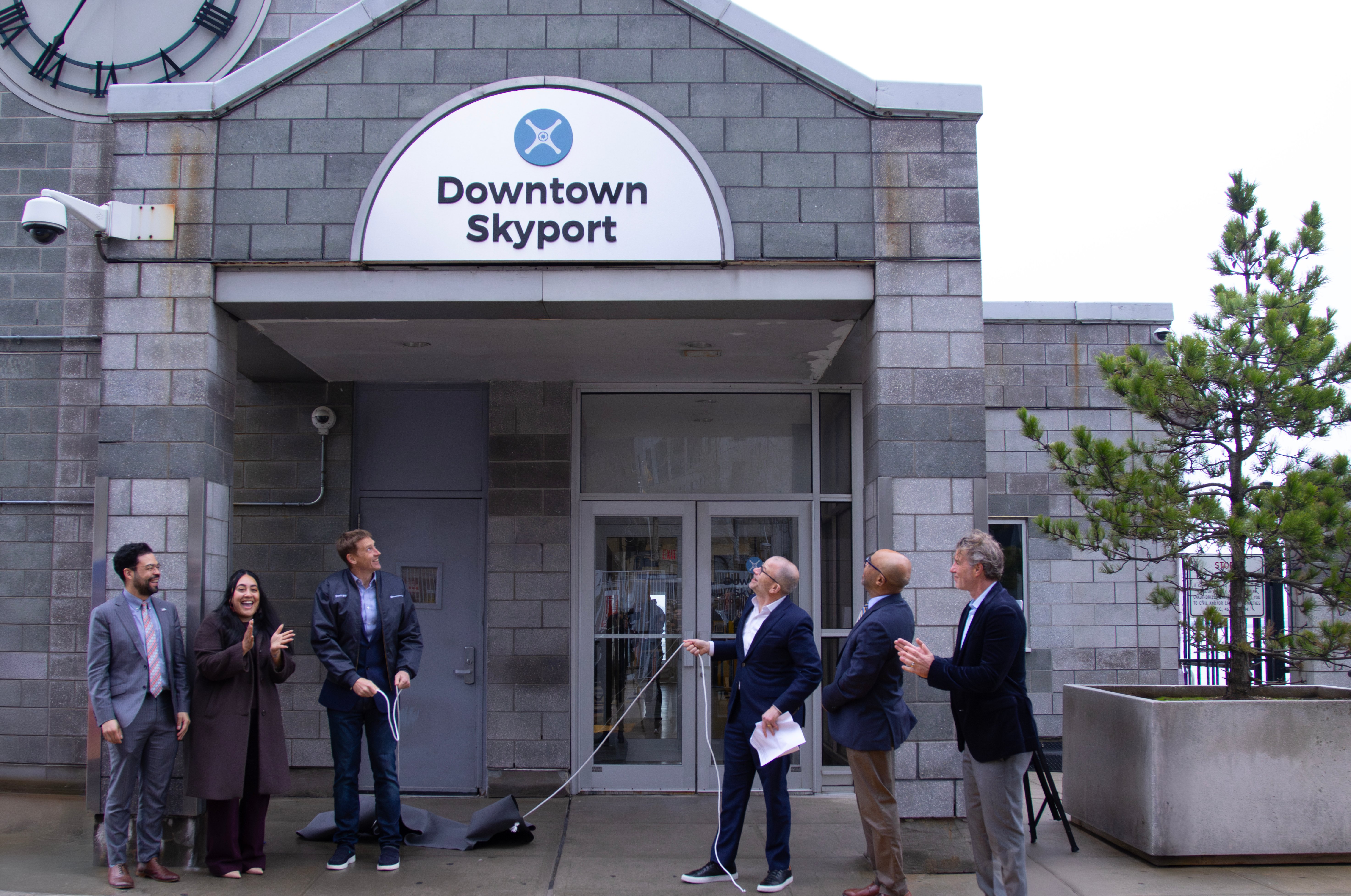 A group of six people, dressed in business attire, stand in front of a building labeled Downtown Skyport. One person is pulling a cord, possibly revealing the sign above the entrance. The setting appears formal and celebratory.