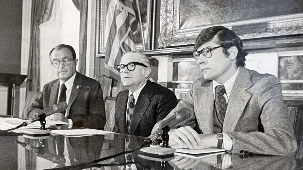 Three men in suits and glasses sit at a table with microphones in front of them, appearing to speak at a formal press conference. An American flag and framed paintings are visible in the background.