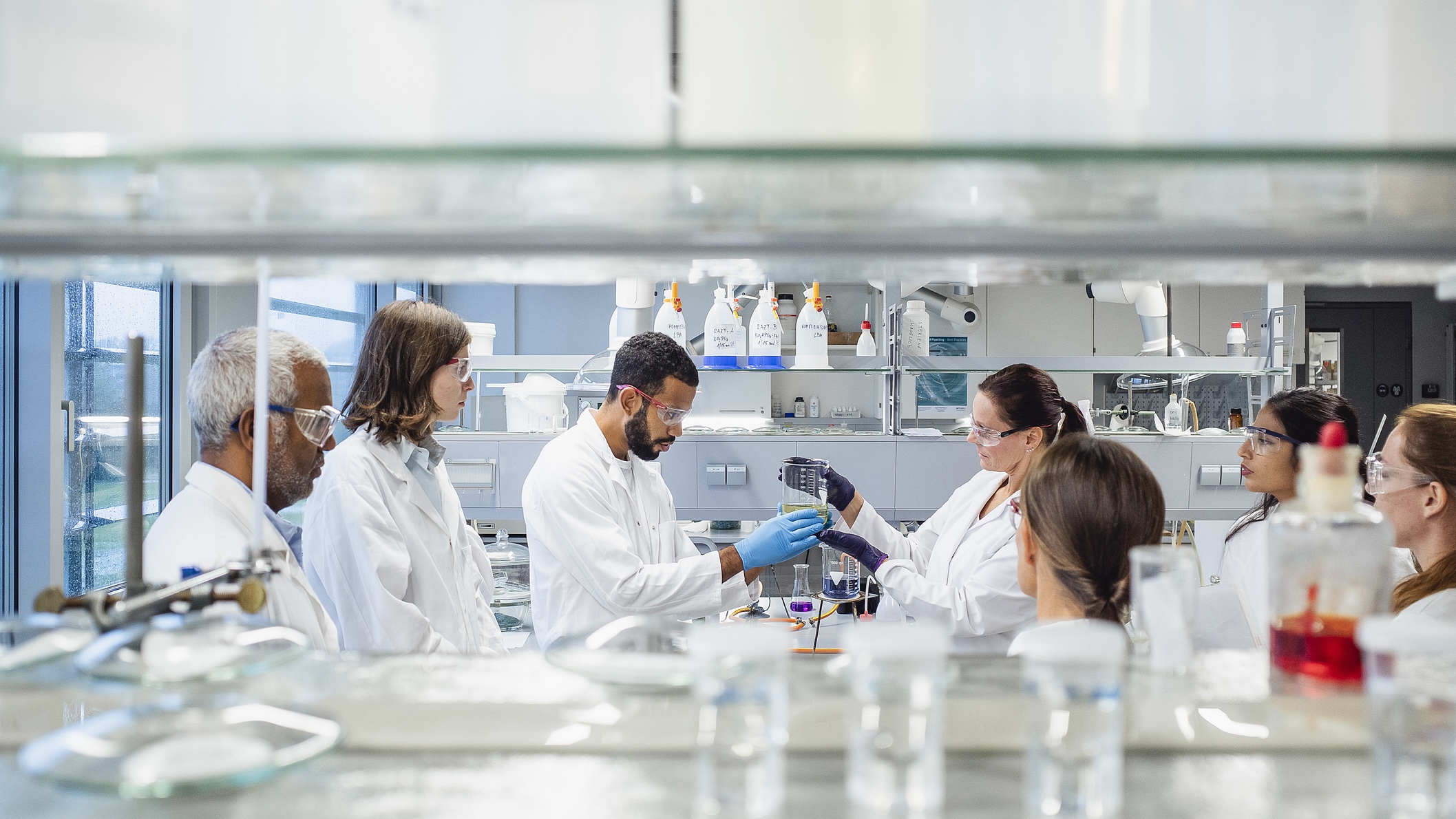 Group of scientists in white lab coats and safety goggles stand around a lab bench, observing and discussing an experiment in a modern laboratory.