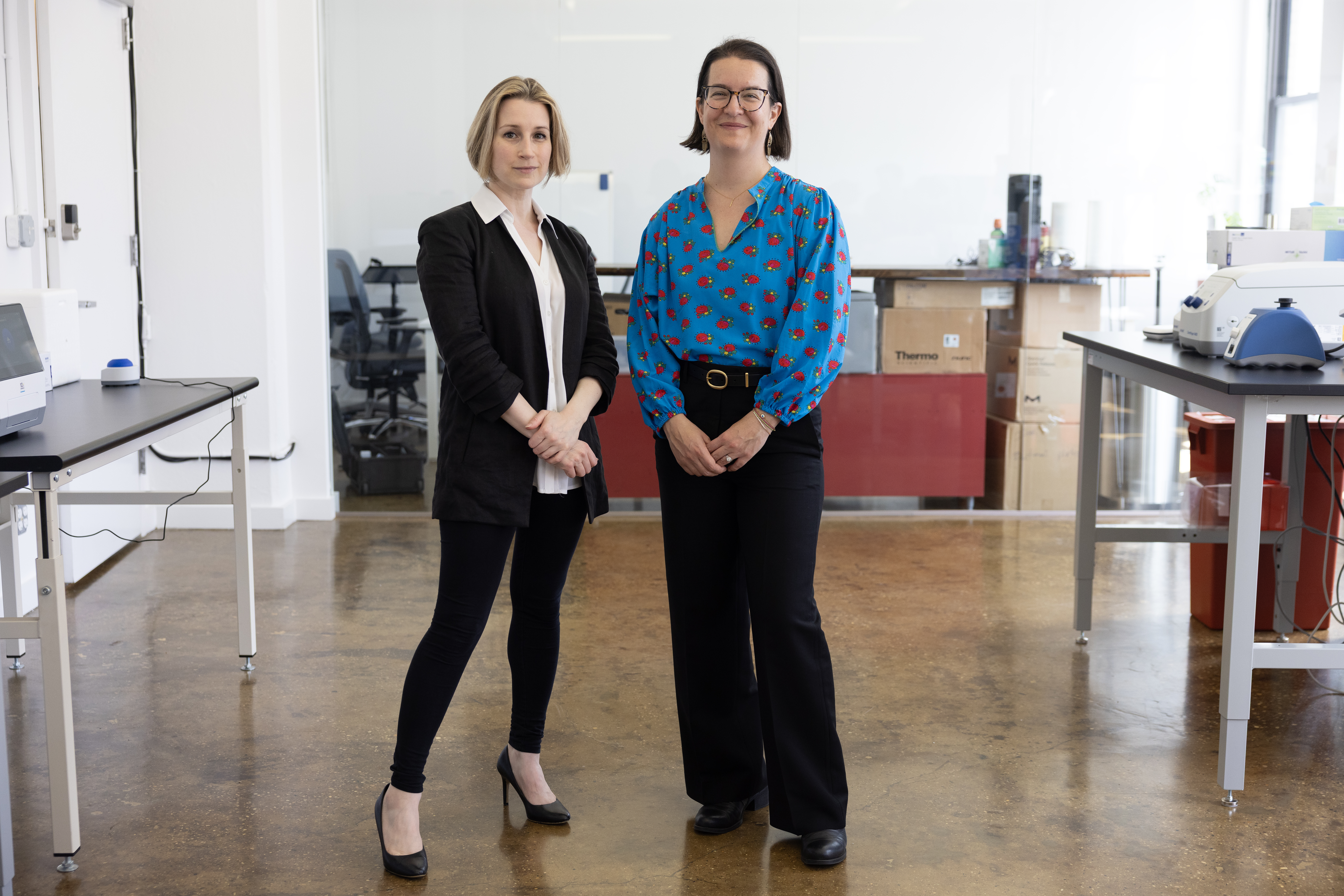 Two women, Cecilia Kushner and Beck Brachman, stand side by side in a modern, bright laboratory. One wears a black suit and heels, the other a blue blouse with a red pattern and black pants. Lab equipment and workstations are visible in the background.