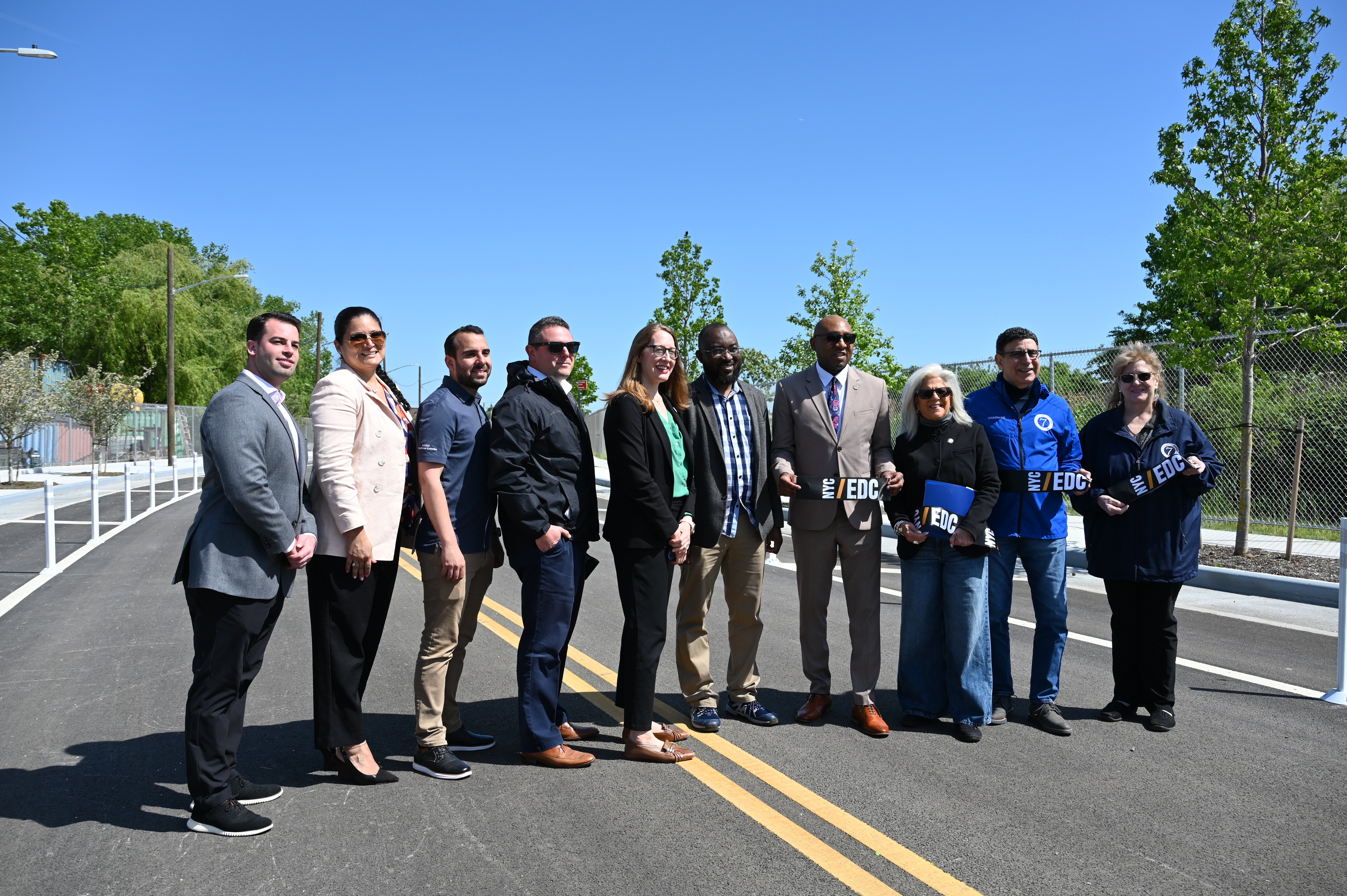 Group of ten people stand together on a newly paved 132nd Street extension in College Point, smiling at the camera. Some are holding a ribbon with the NYCEDC logo on it. Trees and construction equipment are visible in the background under a clear blue sky.