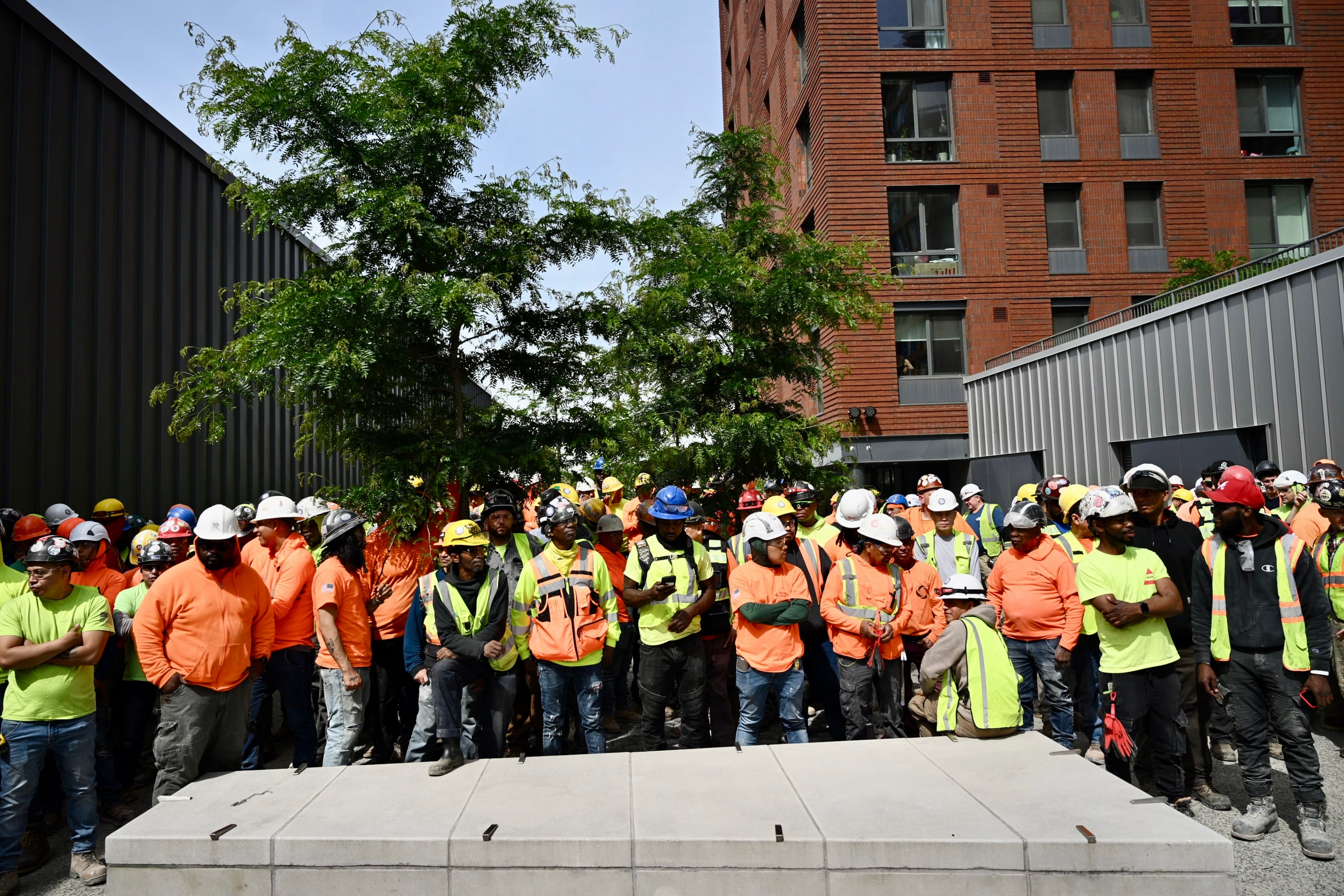 Large group of construction workers wearing safety vests and hard hats stand together outside Hunts Point Peninsula, some with arms crossed, listening attentively. A leafy tree is in the background.