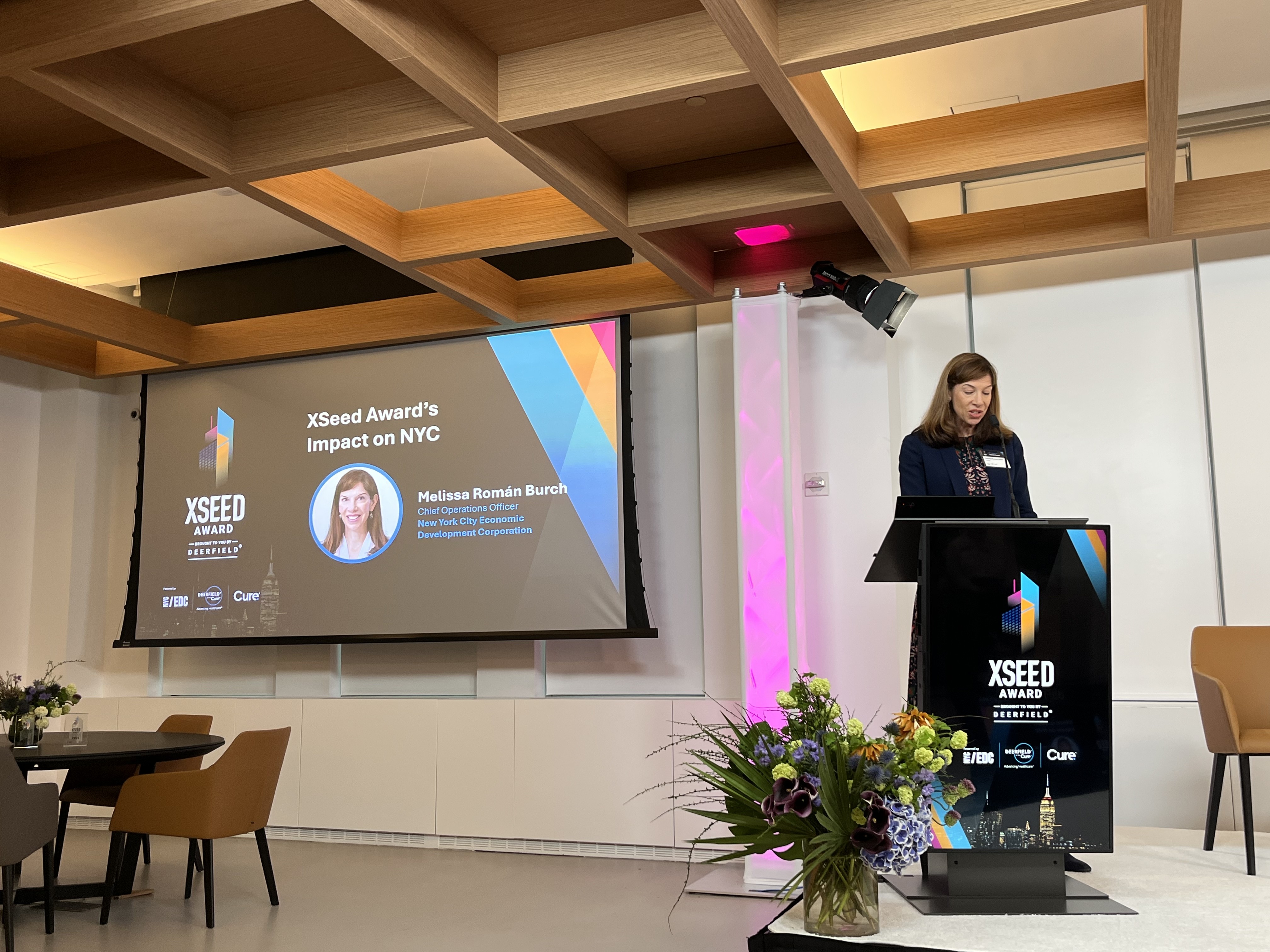 A woman stands at a podium speaking at the XSeed Award event. A large screen behind her displays the title XSeed Award’s Impact on NYC and her name, Melissa Román Burch. Flowers are arranged near the podium.