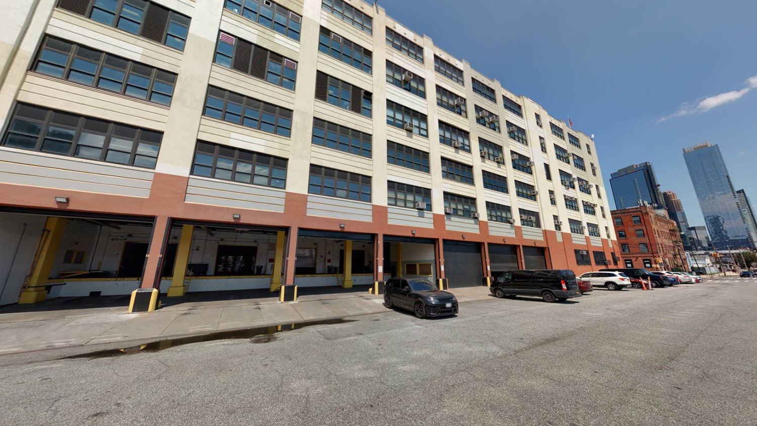 A wide, five-story industrial building with large windows and multiple garage doors at street level. Several cars are parked along the building, and city skyscrapers are visible in the background under a clear sky.