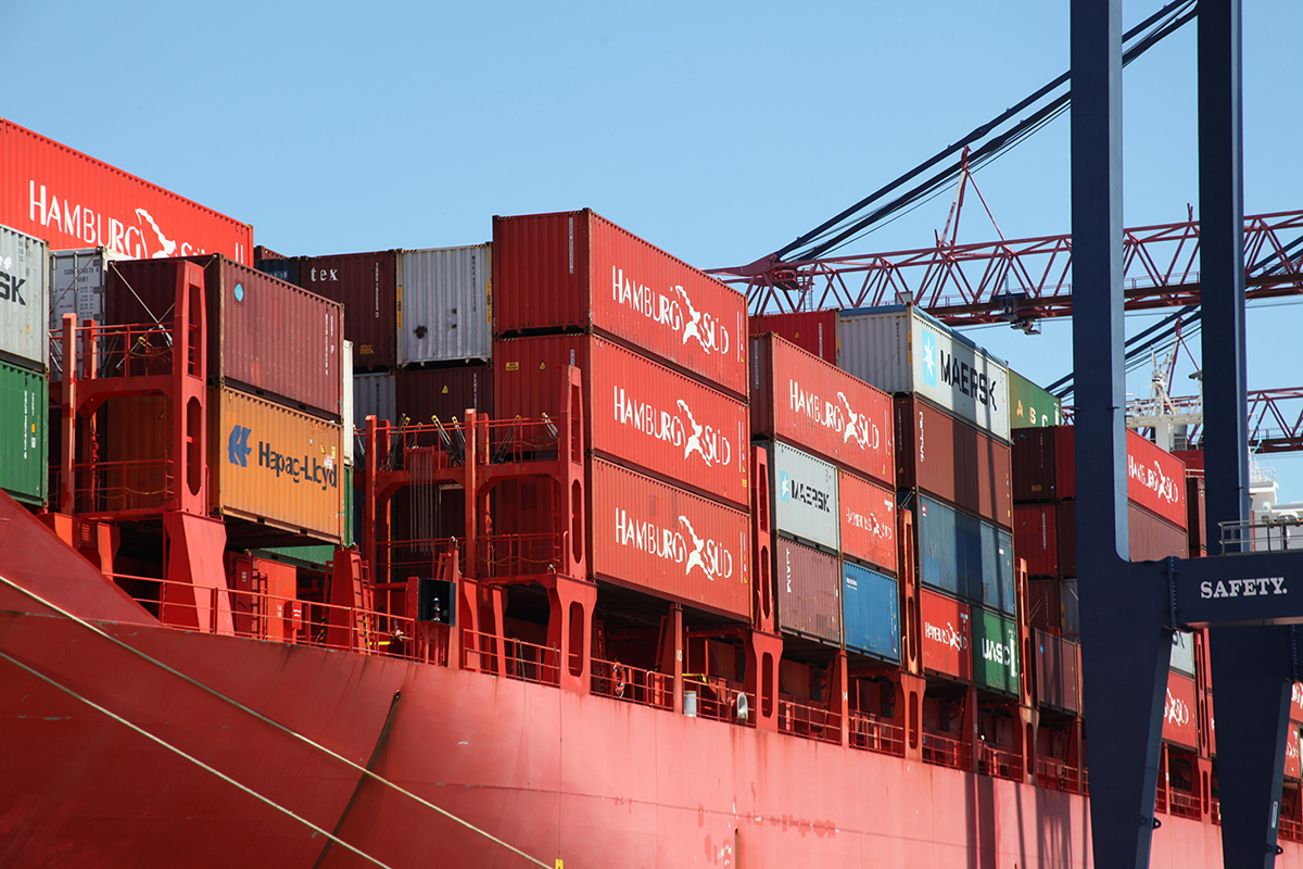 A large red cargo ship loaded with colorful shipping containers is docked at a port, with cranes and blue sky visible in the background.