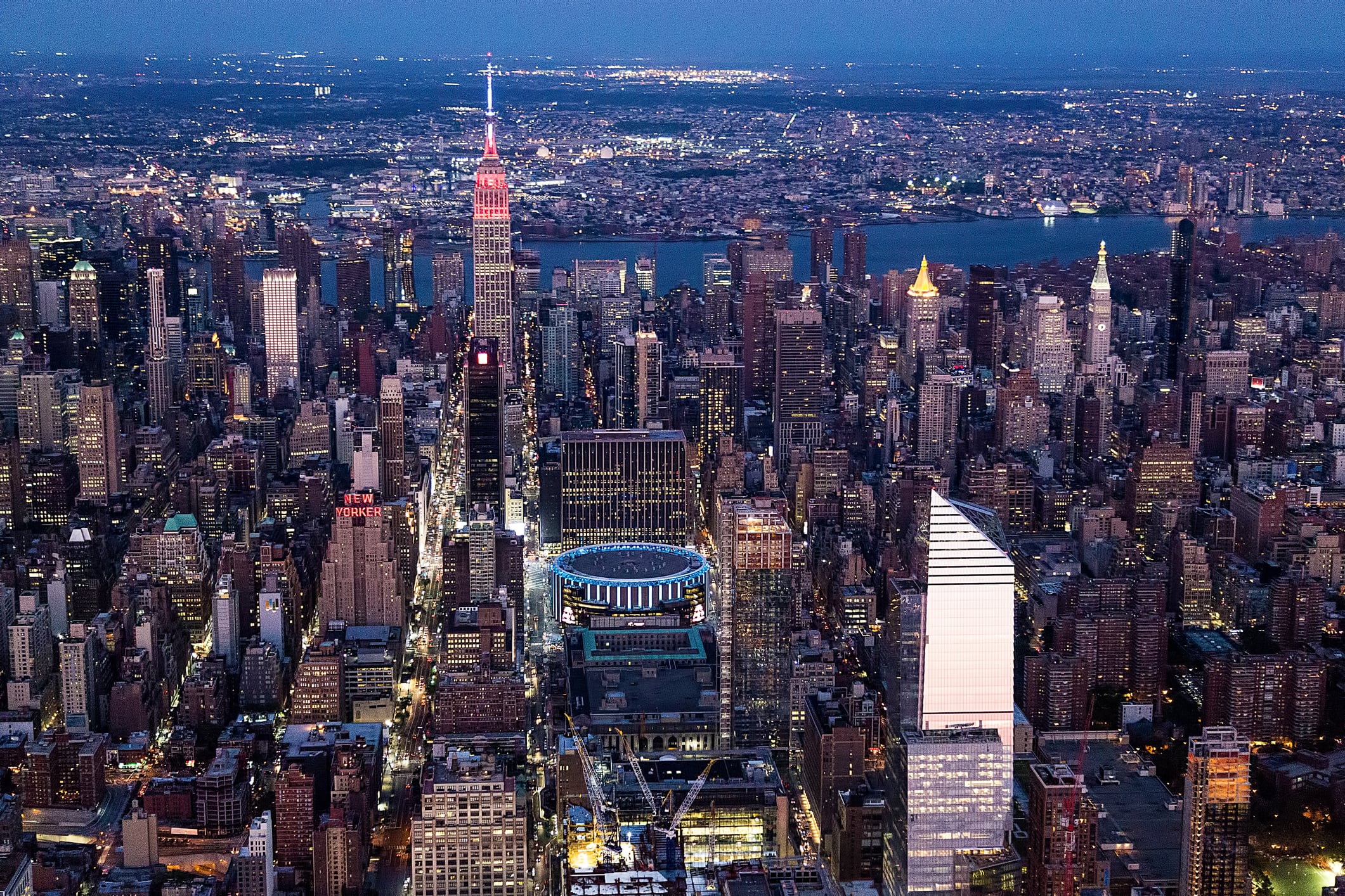 Aerial view of Midtown Manhattan at dusk, featuring the glowing Empire State Building, Madison Square Garden, and a dense cluster of skyscrapers with city lights, set against a backdrop of the Hudson River and distant cityscape.