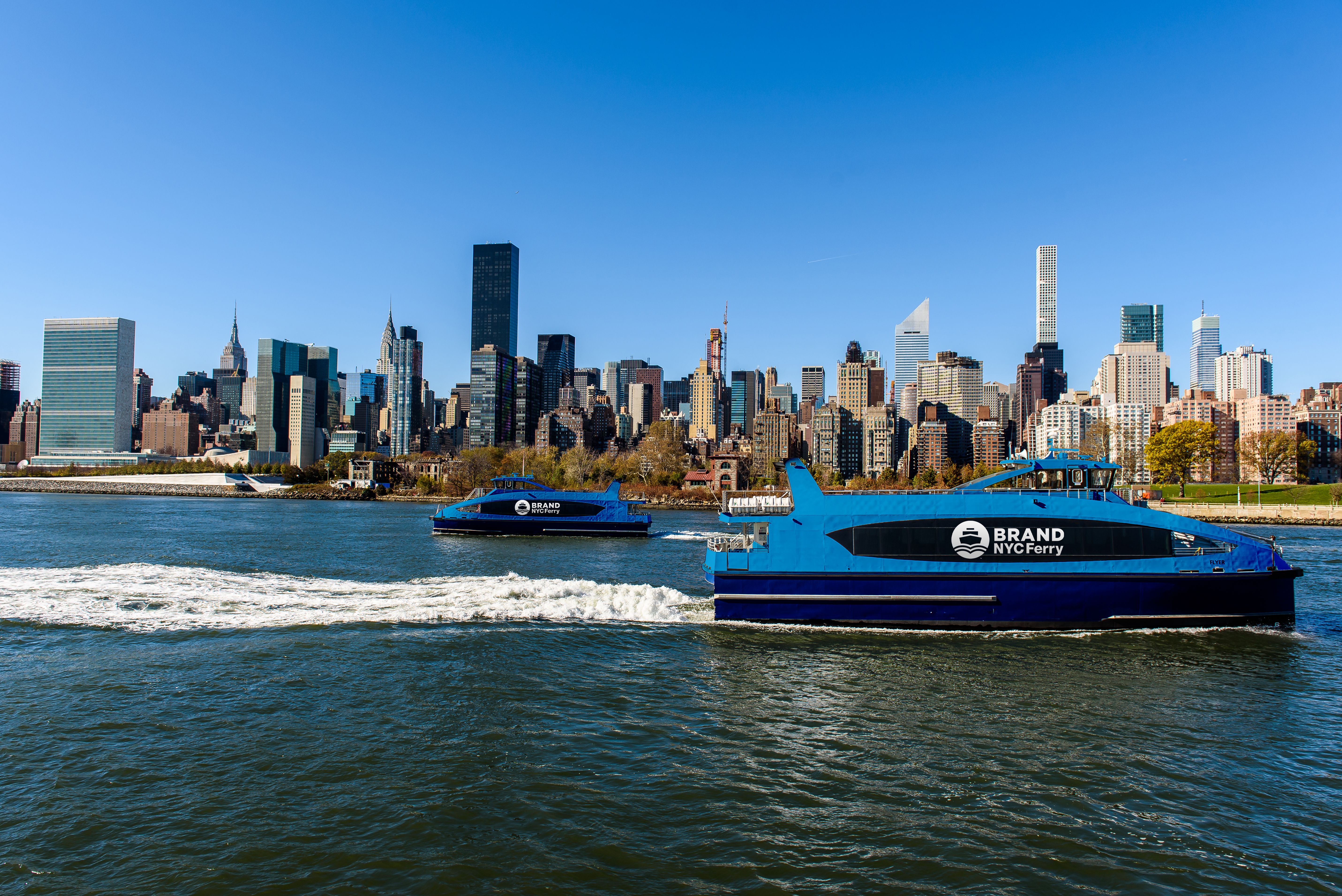 Two blue NYC ferries, each labeled BRAND NYC FERRY, travel on the river. The New York City skyline, including tall skyscrapers and clear blue sky, is in the background.