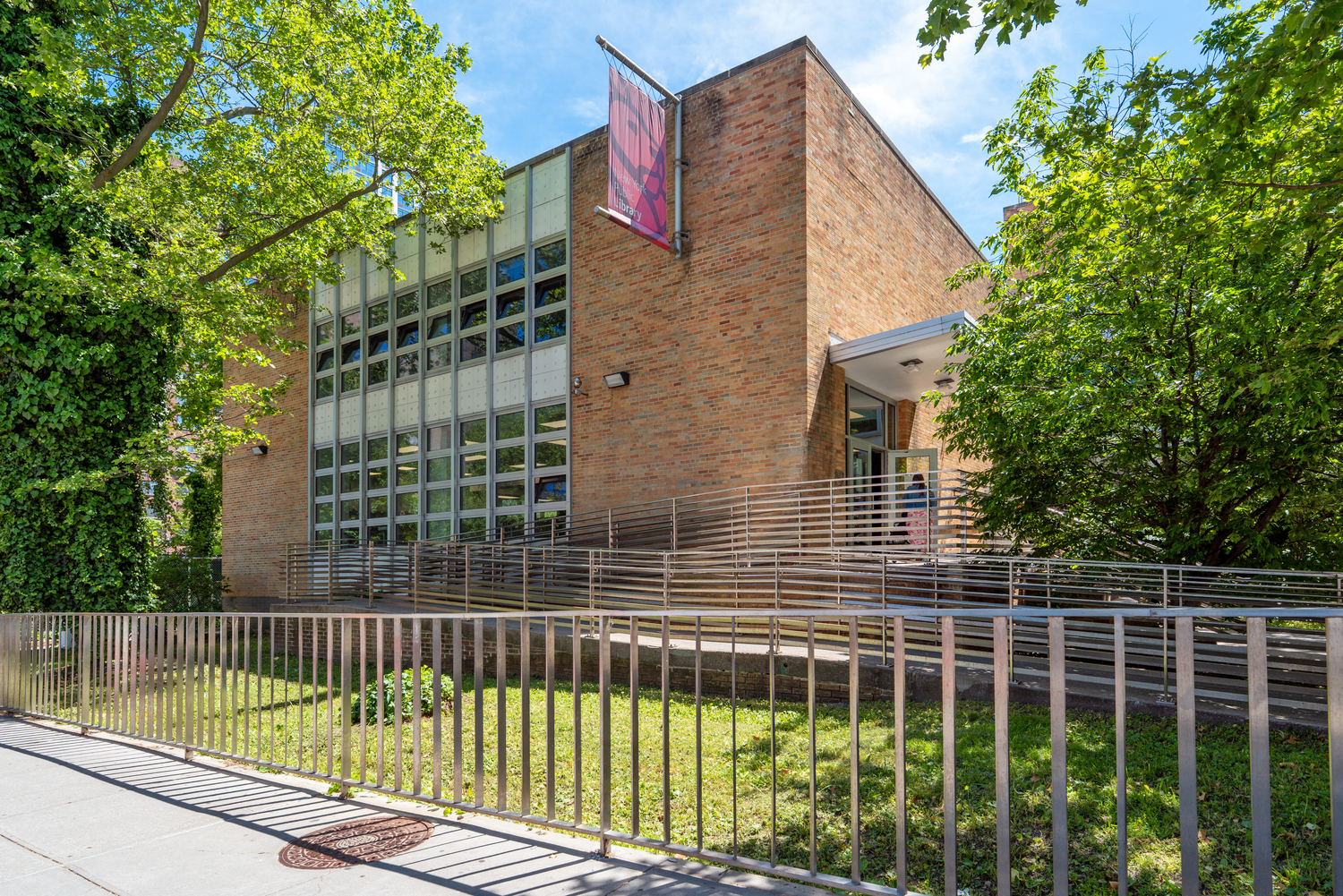 A modern brick building with large grid-like windows, a sidewalk with a metal fence in front, and leafy green trees surrounding the entrance. A red New York Public Library banner hangs near the entrance on a sunny day.