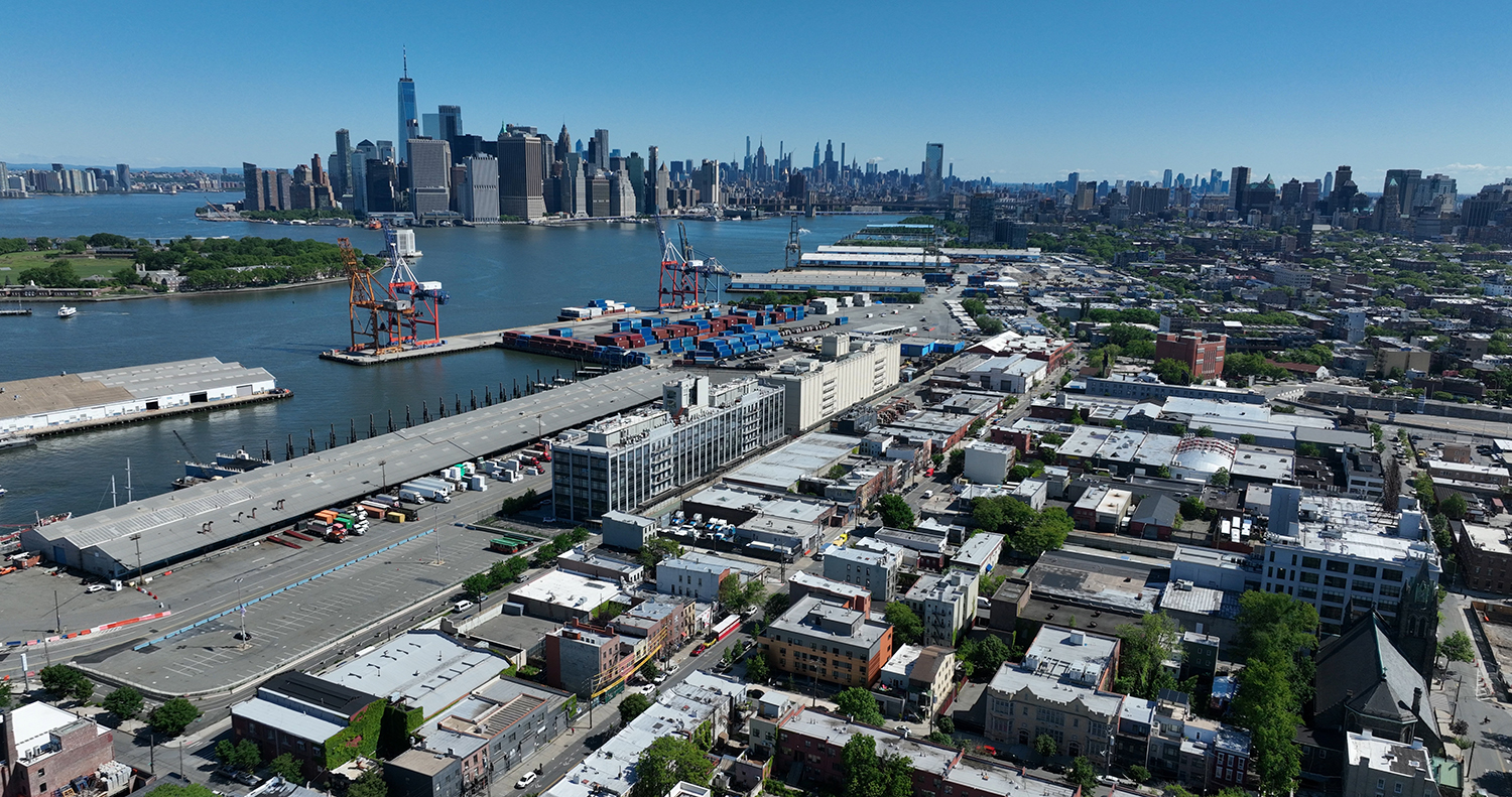 Aerial view of Brooklyn Marine Terminal with shipping containers and cranes beside an urban area; the Manhattan skyline and waterfront are visible across the river under a clear blue sky.