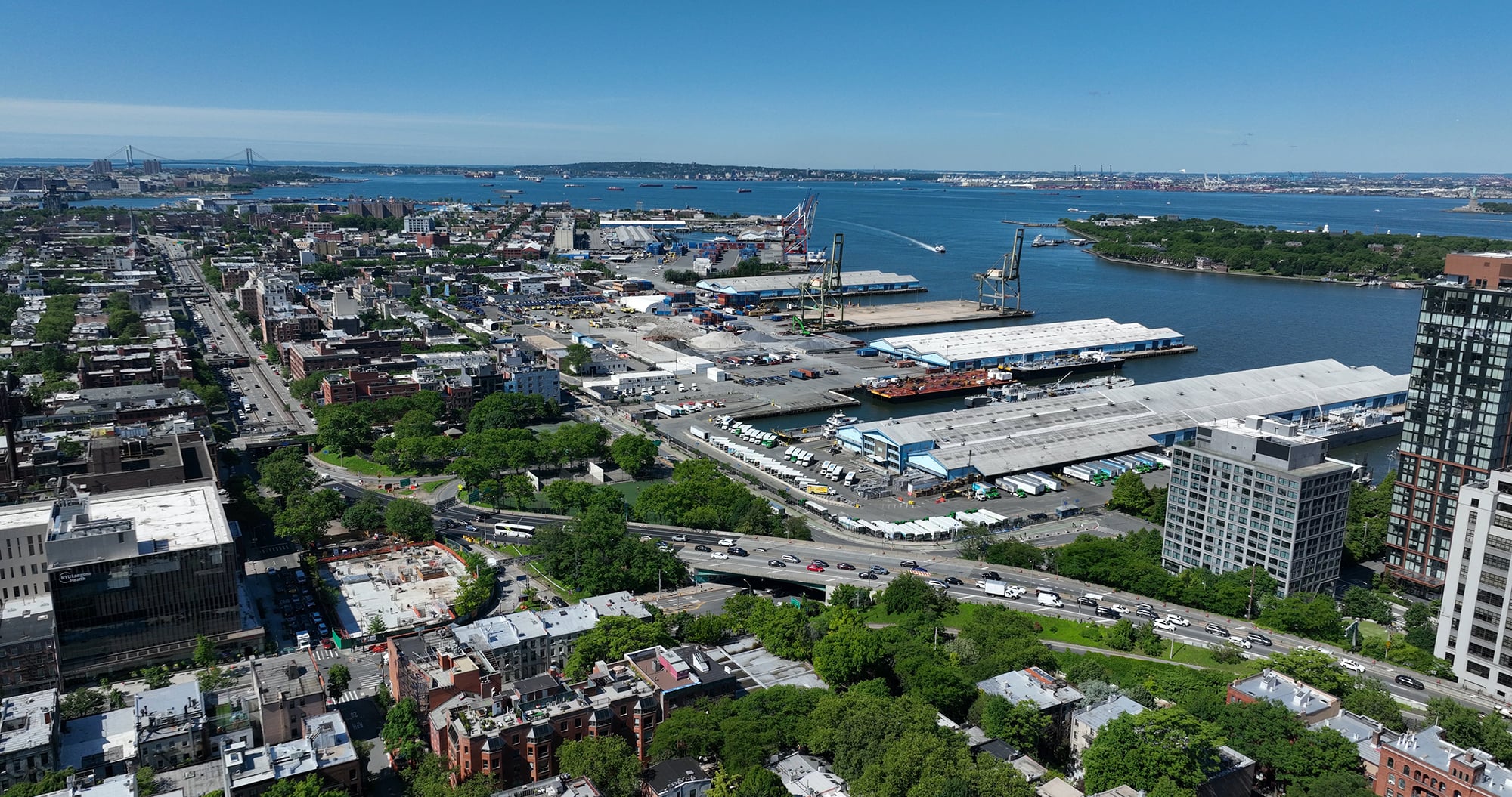 Aerial view of the Brooklyn Marine Terminal with industrial buildings, shipping docks, highways, green spaces, and a river; bridges and islands are visible in the distance under a clear blue sky.