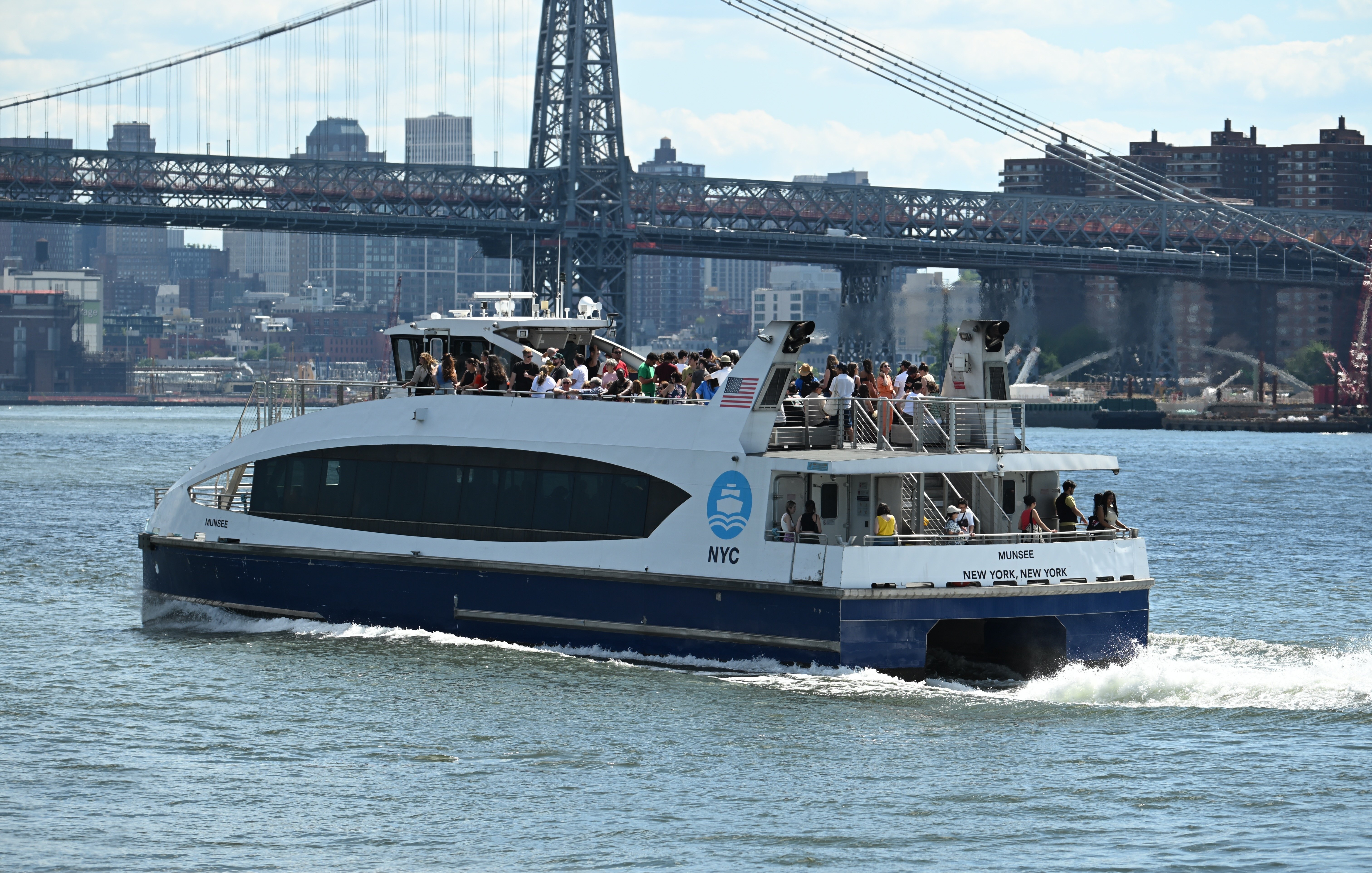 A white and blue NYC ferry with passengers on board travels along the water, with a New York City skyline, buildings, and a bridge visible in the background. Photo courtesy of John Vital.