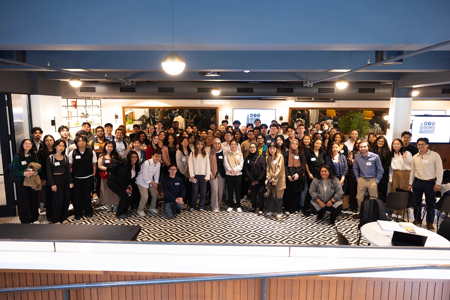 NYC Startup Internship program participants pose for a group photo indoors, many wearing name tags and business attire, standing on a black-and-white patterned floor in a spacious, modern room.