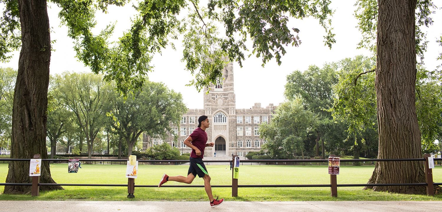 A man in athletic wear jogs on the Fordham University campus along a path lined with trees, with a large, historic stone building and green lawn in the background on a bright, clear day.