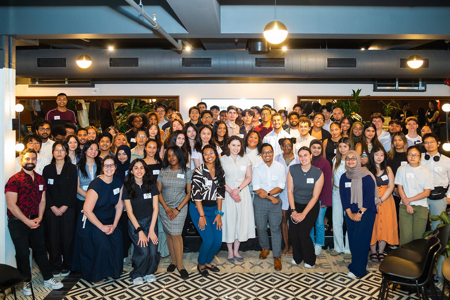 A large, diverse group of people poses together and smiles for a group photo in a brightly lit, modern indoor space with patterned flooring and plants in the background.