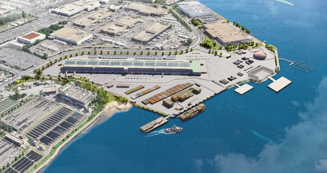 Rendering of the proposed Hunts Point Marine Terminal. An aerial view of an industrial waterfront facility with warehouses, shipping containers, parked vehicles, a pier, and a boat on blue water, surrounded by urban buildings and roads. Rendering courtesy of the City of New York.