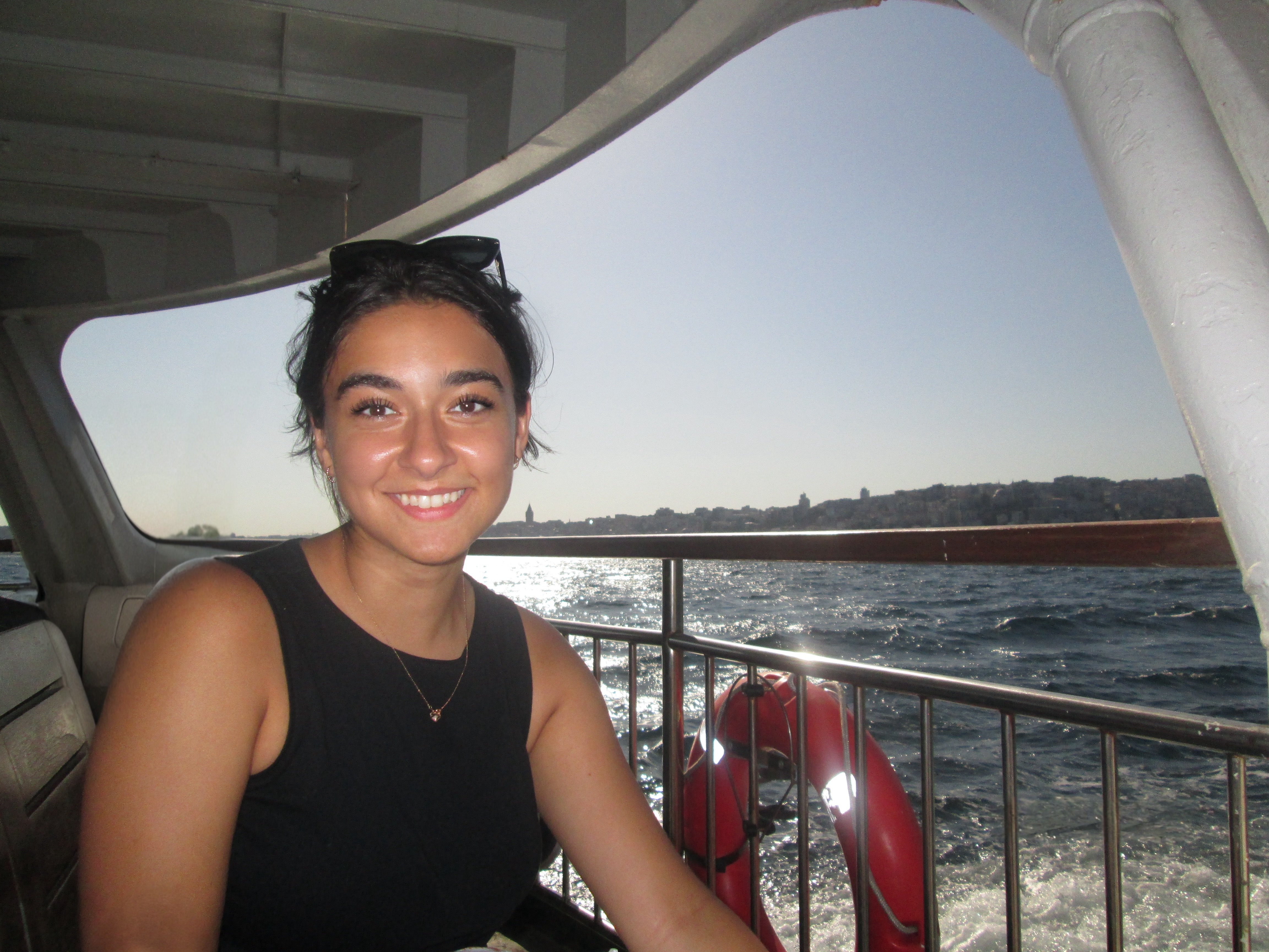 Woman with dark hair and a black sleeveless top, Leyla Arcasoy, smiles while sitting on a boat. The sun is shining brightly over the water in the background, and a cityscape is visible on the horizon.