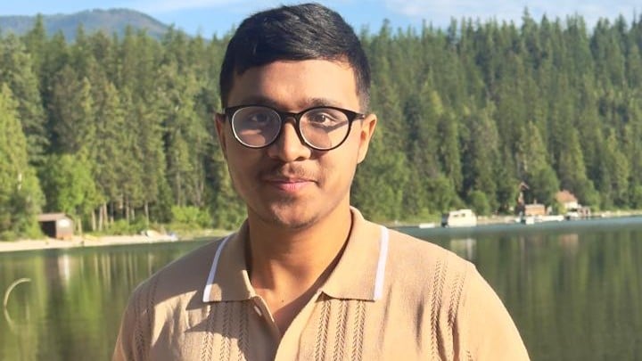 Debashrestha Nandi wearing glasses and a beige shirt stands by a lake with a forest and mountains in the background on a sunny day.