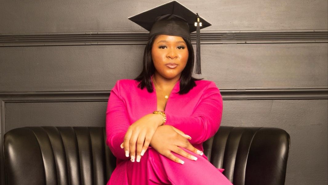 Femi Jordan wearing a bright pink suit and graduation cap, sits confidently on a black leather couch against a dark wall, looking directly at the camera with her hands crossed on her knee.