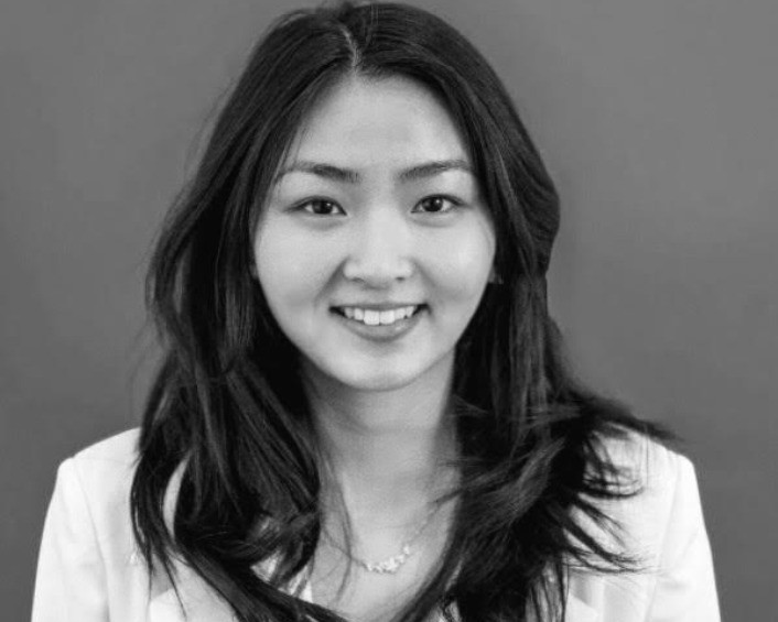 Woman with long dark hair, Justine Cheng, smiles at the camera, wearing a light-colored blazer and necklace, against a plain background. The image is in black and white.