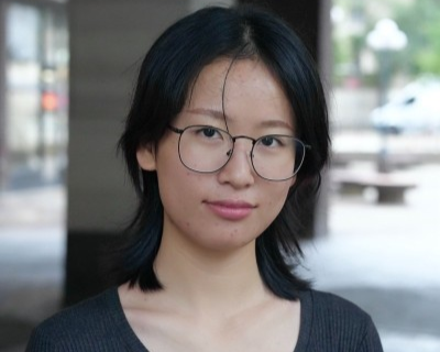 Kelly Lin with straight black hair, wearing round glasses and a dark top, looks at the camera with a slight smile. The background is blurred, showing an outdoor urban setting.