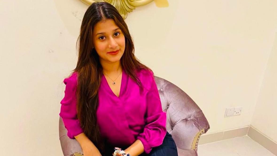 A woman with long brown hair, Samrin Alam, wearing a bright pink blouse and dark pants, sits on a light purple tufted chair in a room with cream-colored walls and a decorative mirror behind her.