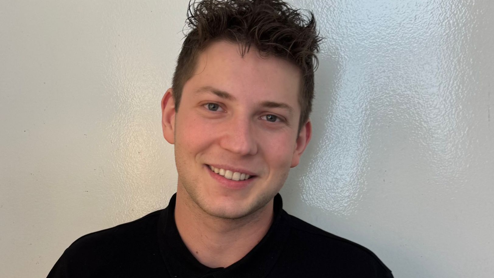 Alexandru Daniel Soroiu with short, curly brown hair and a friendly smile stands against a plain light-colored wall, wearing a black shirt.