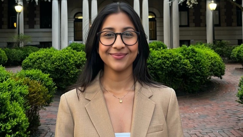 Anushka Khetawwat with long dark hair and glasses, wearing a beige blazer and a necklace, stands smiling in front of a building with columns and green bushes.
